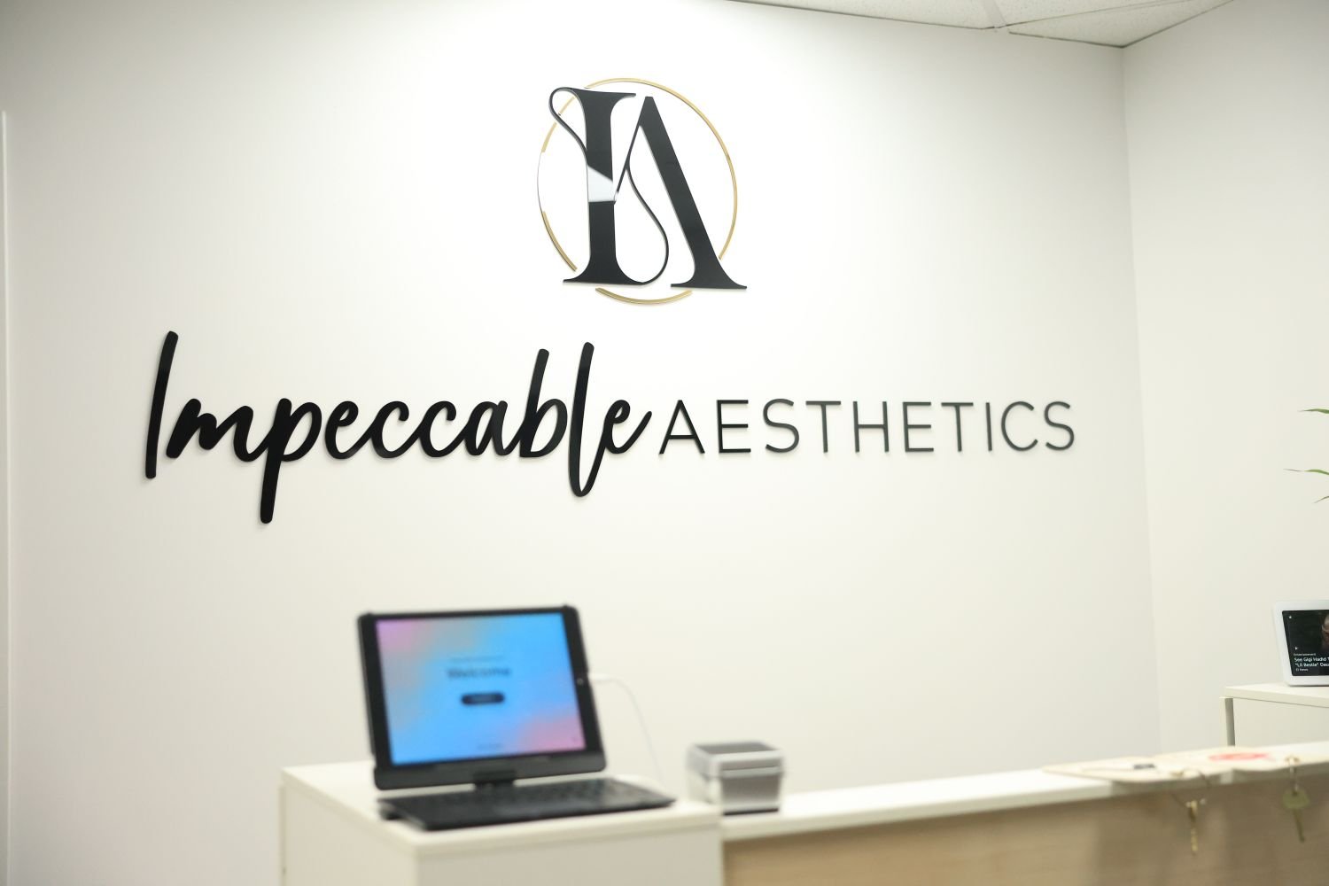 Reception area of Impeccable Aesthetics with a white wall, black logo with initials 'IA', and the business name 'Impeccable Aesthetics' in black cursive and uppercase letters.