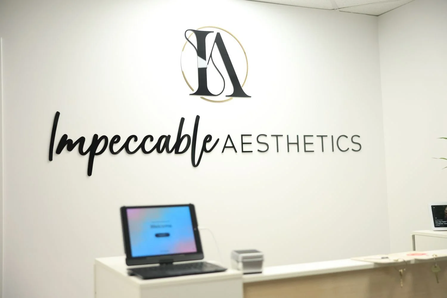 Reception area of Impeccable Aesthetics with a white wall, black logo with initials 'IA', and the business name 'Impeccable Aesthetics' in black cursive and uppercase letters.