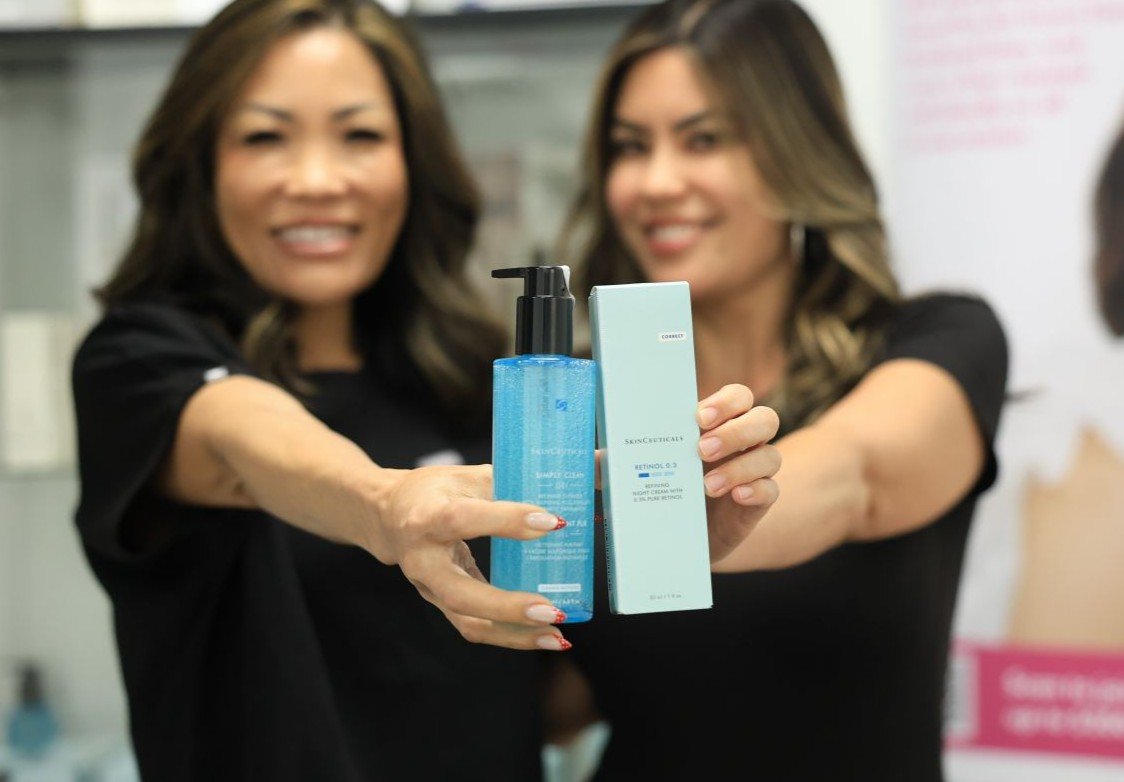 Two women in black shirts holding skincare products, with one product in clear view, in a store or clinic.