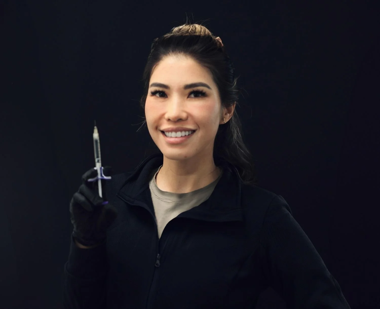 A woman smiling and holding a syringe in her right hand, wearing black gloves, a black jacket, and light-colored shirt against a dark background.