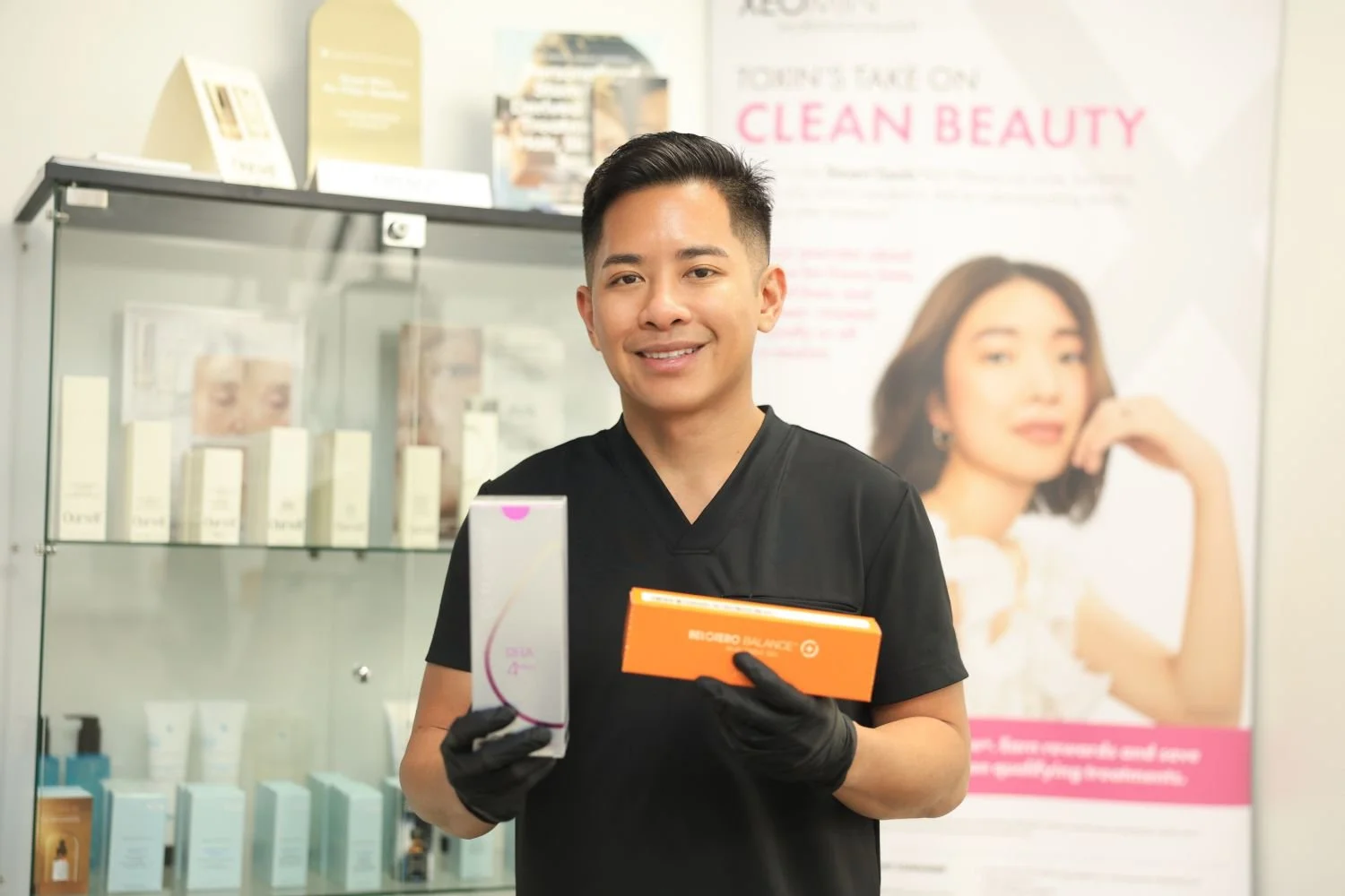 A smiling man in black medical scrubs and black gloves holding skincare product boxes in a store with beauty products and a promotional poster in the background.