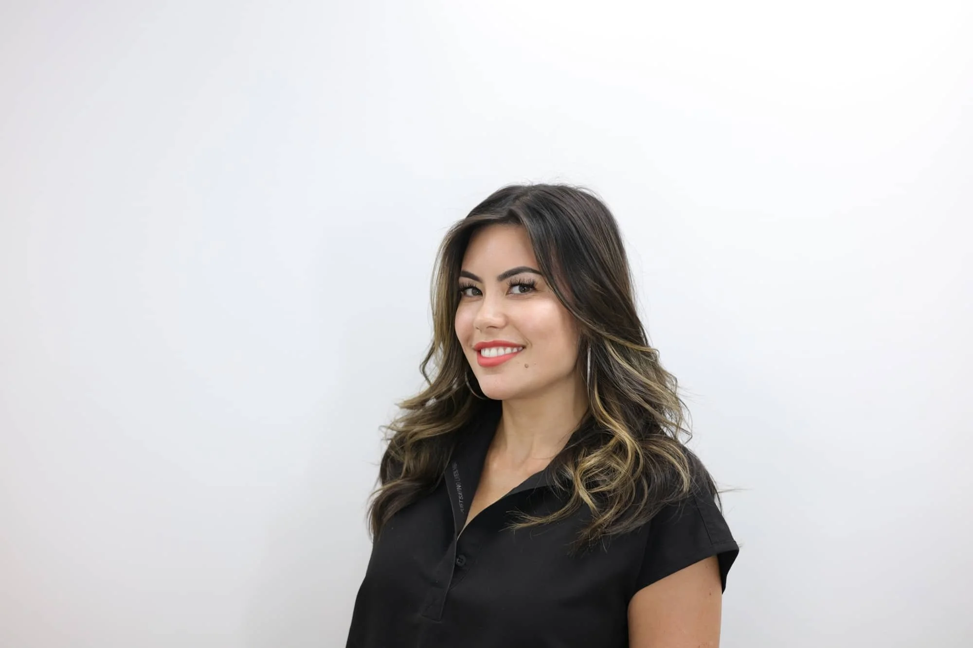 A woman with wavy, shoulder-length brunette hair, wearing a black collared shirt, smiling with teeth showing, standing against a plain white background.