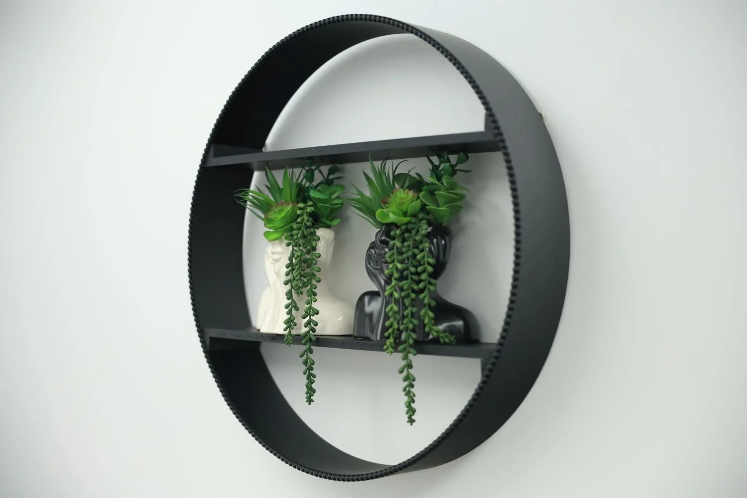 Round black wall shelf with two compartments holding white and black decorative vases with green succulent plants.