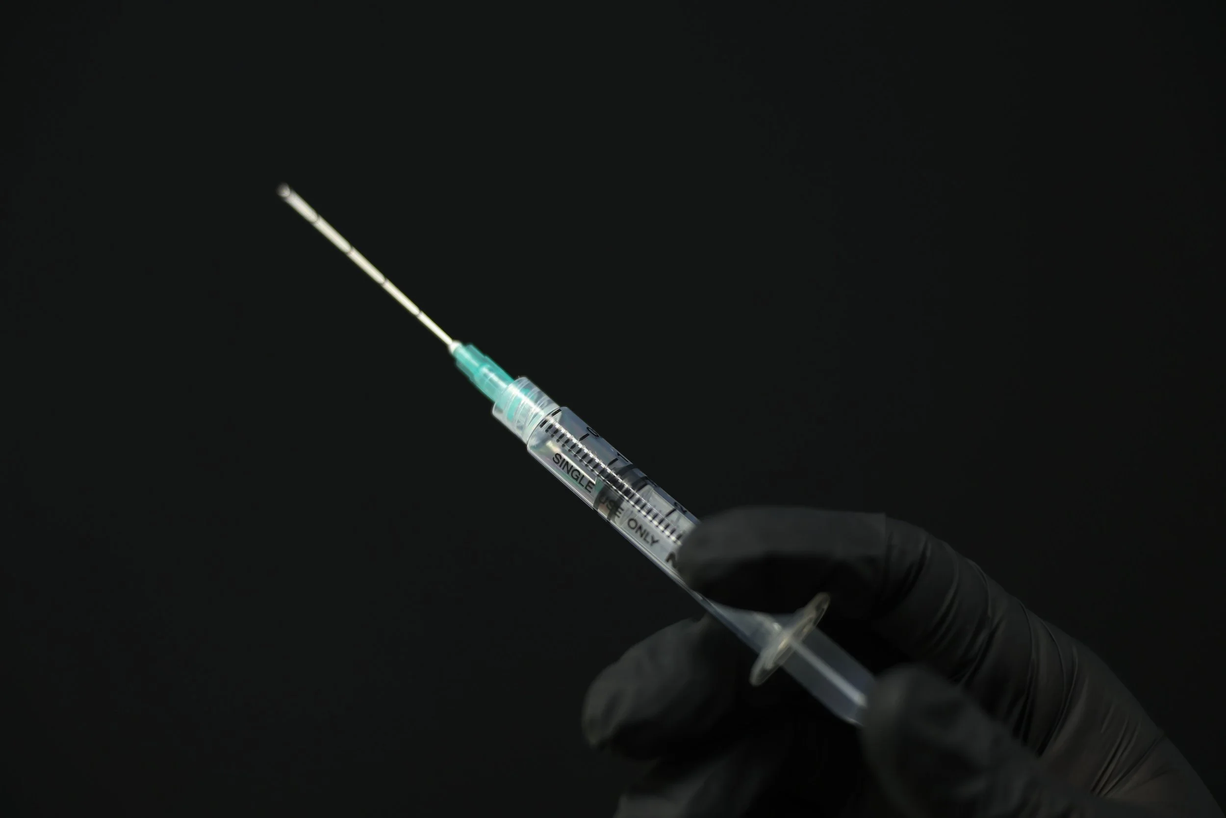 A gloved hand holding a syringe filled with a clear liquid against a black background.