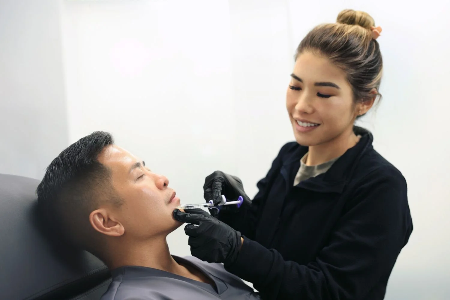 A woman with a bun hairstyle administering a cosmetic injection to a man lying back in a chair, with a white wall in the background.