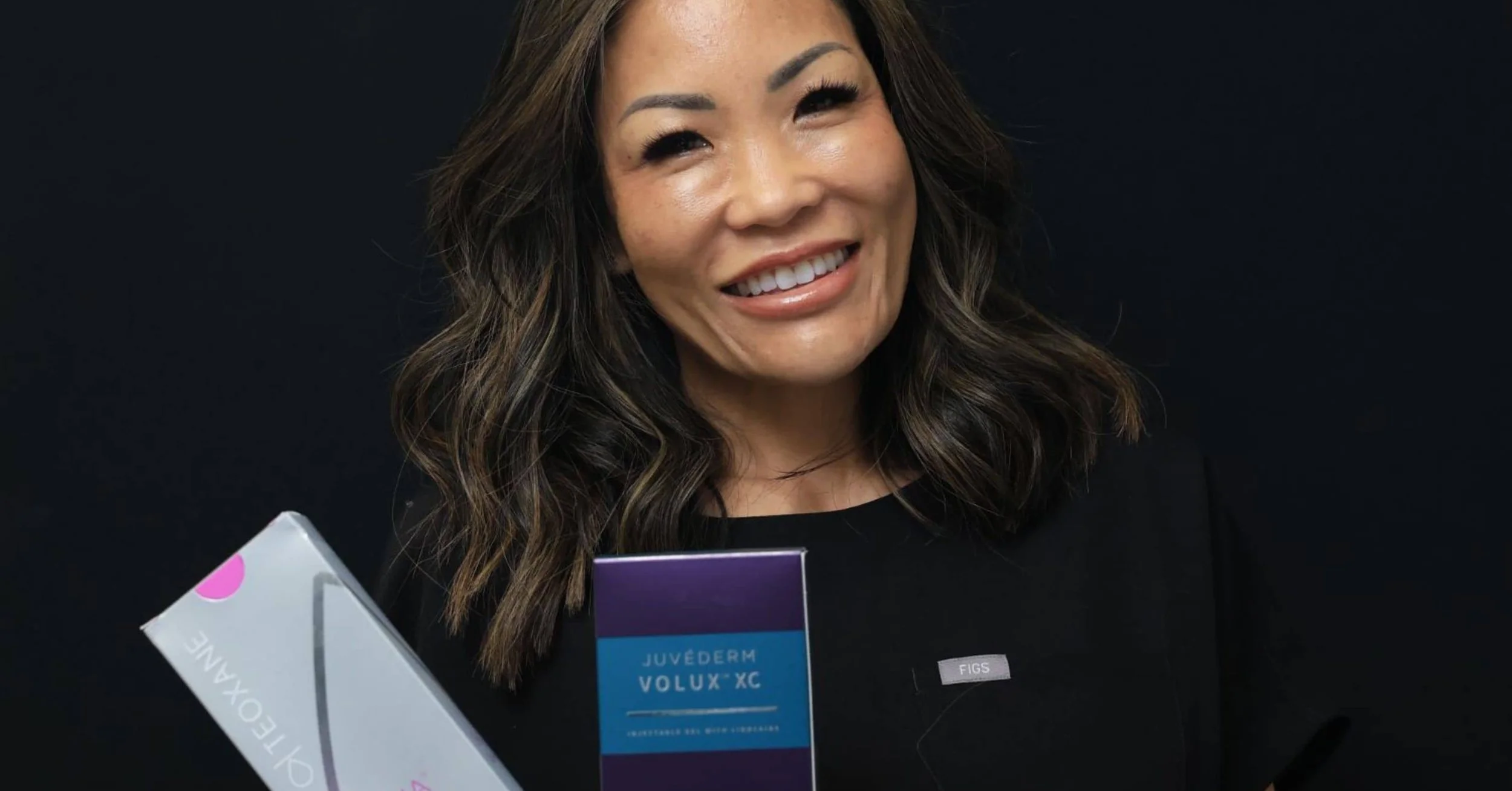 Smiling aesthetic injector holding Teoxane and Juvéderm Volux XC filler boxes used in filler correction treatment.