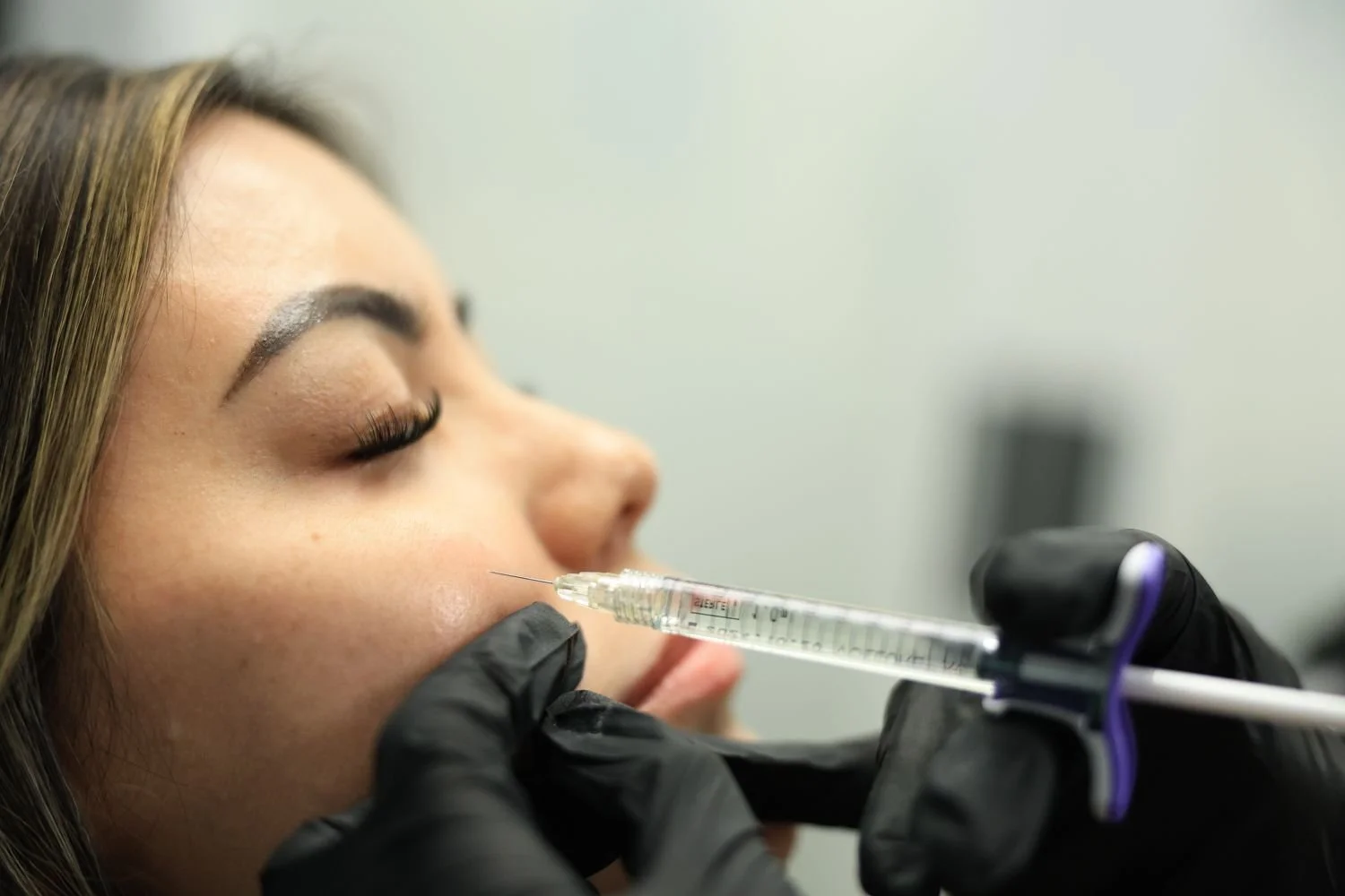 A woman receiving a cosmetic injection in her cheek from a medical professional wearing black gloves.