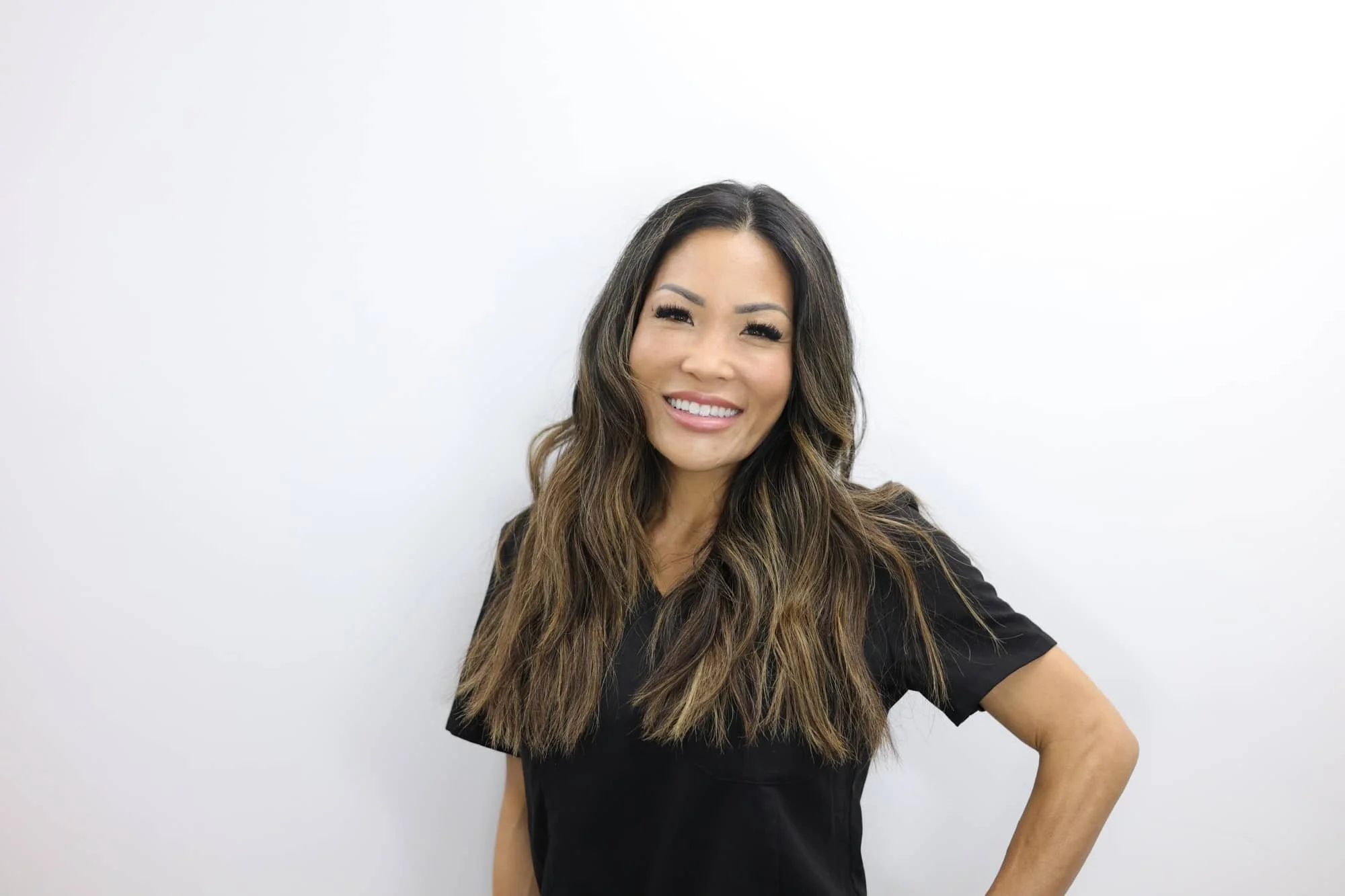 A woman with long wavy brown hair, smiling, wearing a black shirt, standing against a plain white background.