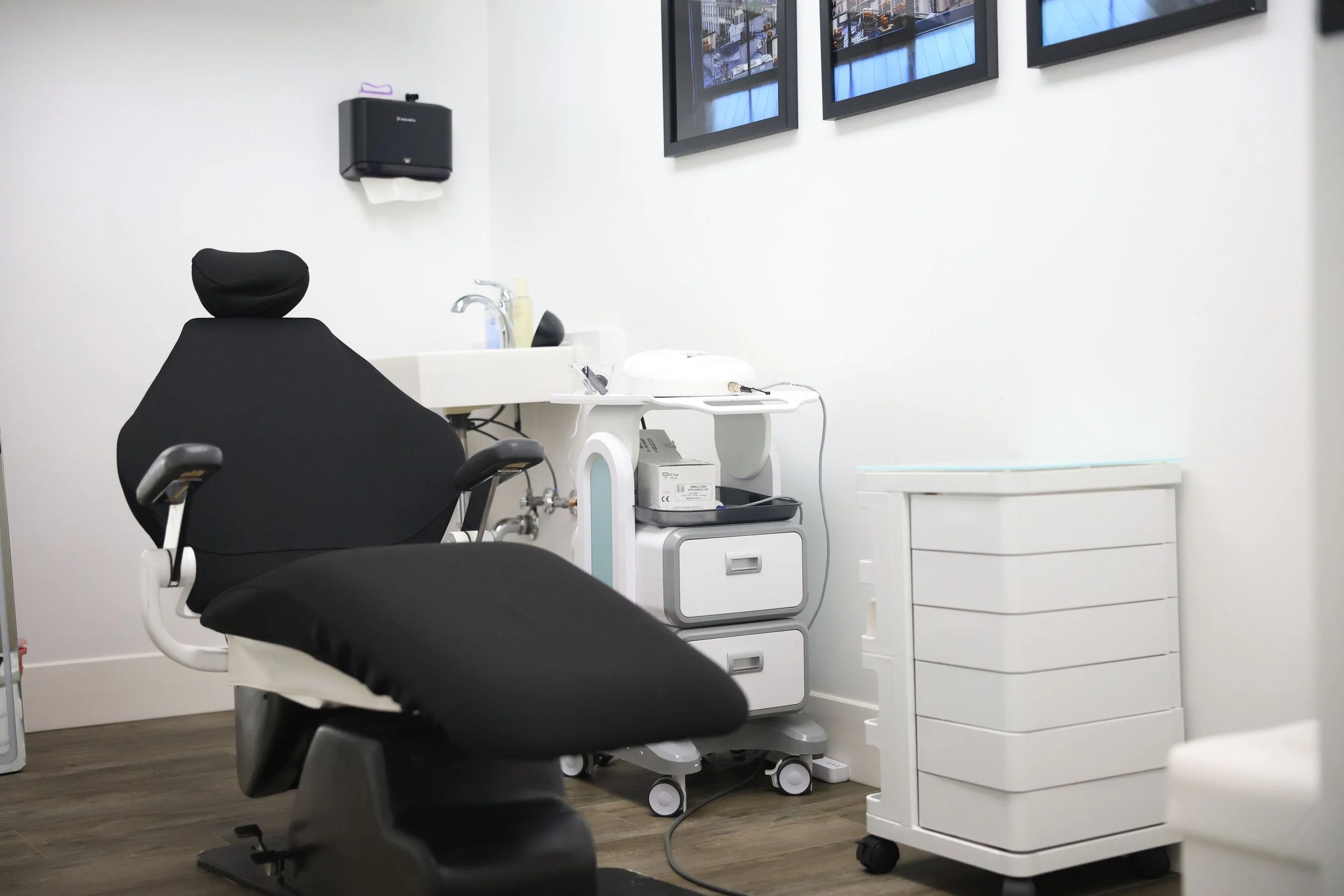 A medical examination room with a black dental chair, a white cabinet with medical equipment, and framed images on the wall.