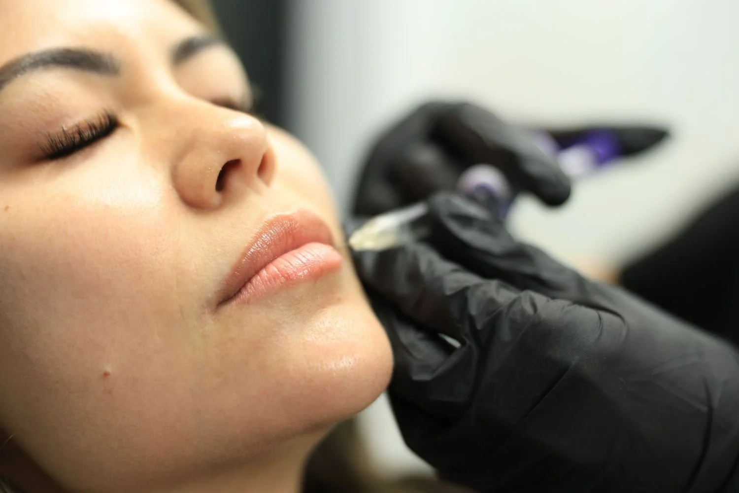 A woman receiving a cosmetic tattoo or microblading procedure on her eyebrows by a technician wearing black gloves.