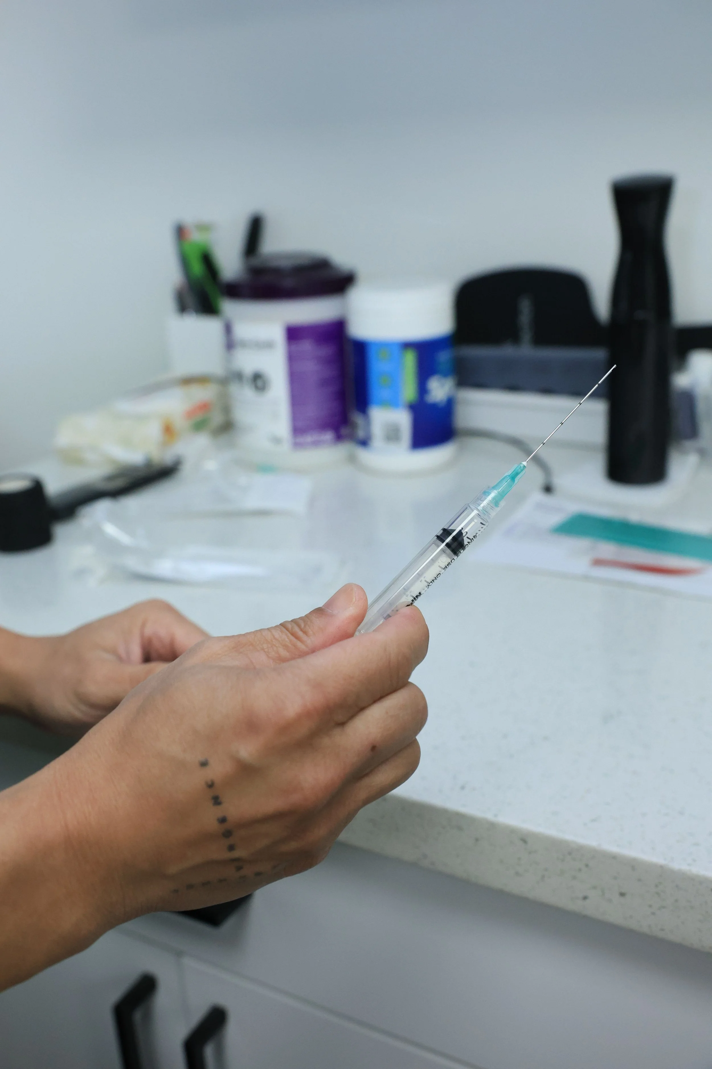 A person holding a syringe in a medical setting, with various medical supplies on a countertop in the background.