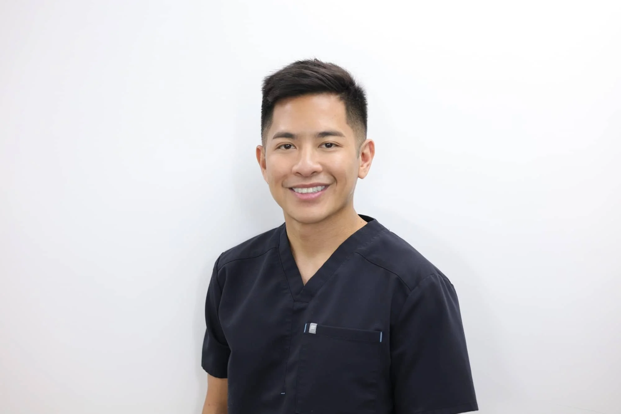 A young man smiling, wearing dark medical scrubs, standing against a plain white wall.