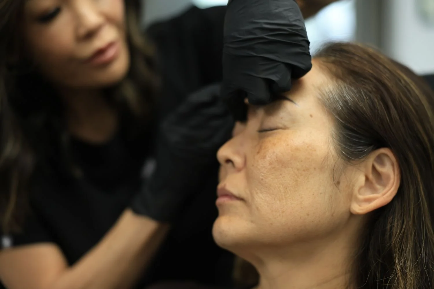 A woman with makeup, lying with eyes closed, receiving a cosmetic procedure on her forehead from a technician wearing black gloves.