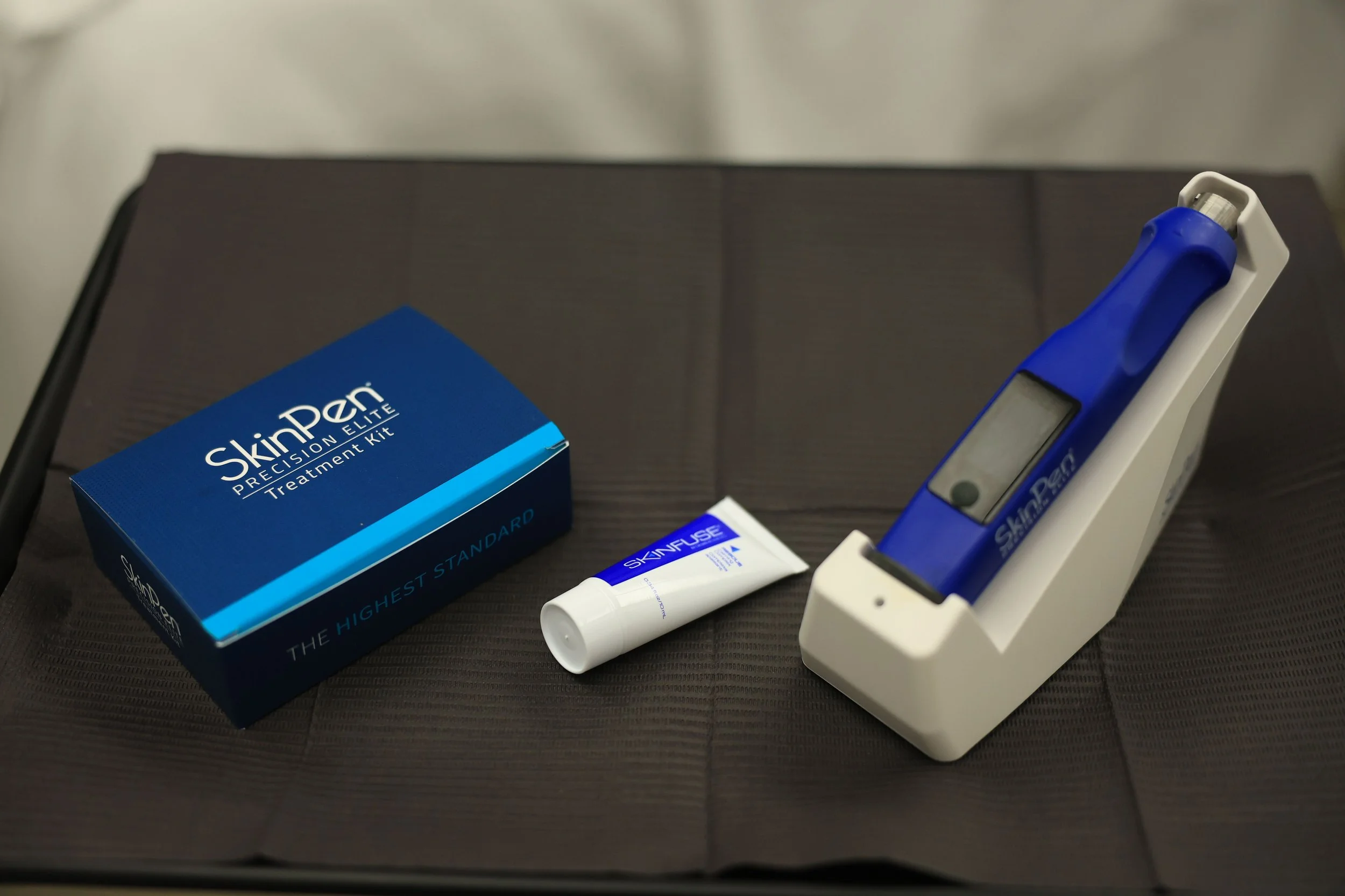 SkinPen Precision Elite treatment kit, including a blue box labeled SkinPen, a small tube of SkinFuse, and a blue and white SkinPen device in a charging stand