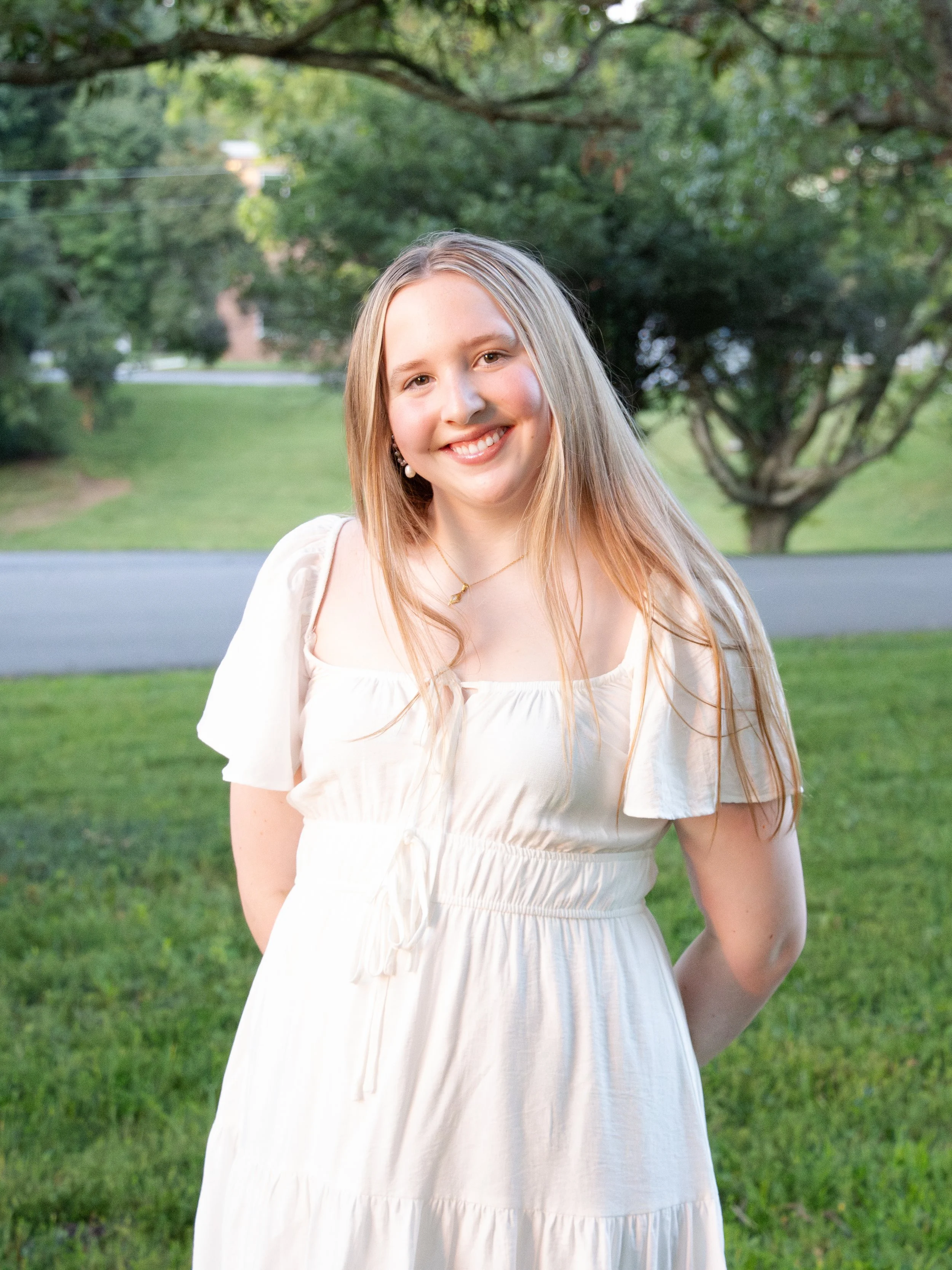 A young woman with long blonde hair and a white dress standing outside in a grassy area with trees and a road in the background, smiling at the camera.