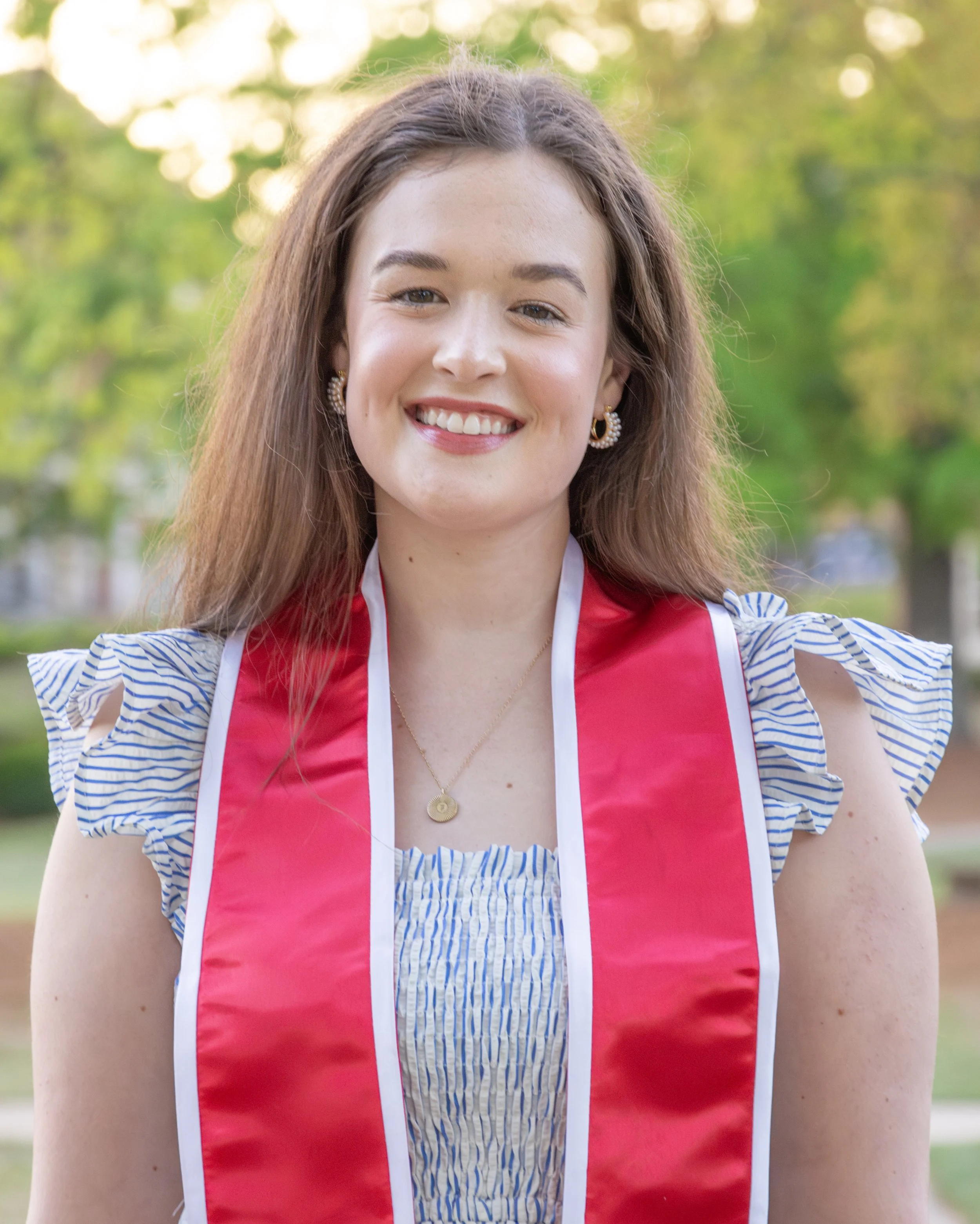 A young woman smiling outdoors with a green, leafy background, wearing a red graduation sash, a striped dress with ruffled sleeves, and gold jewelry.