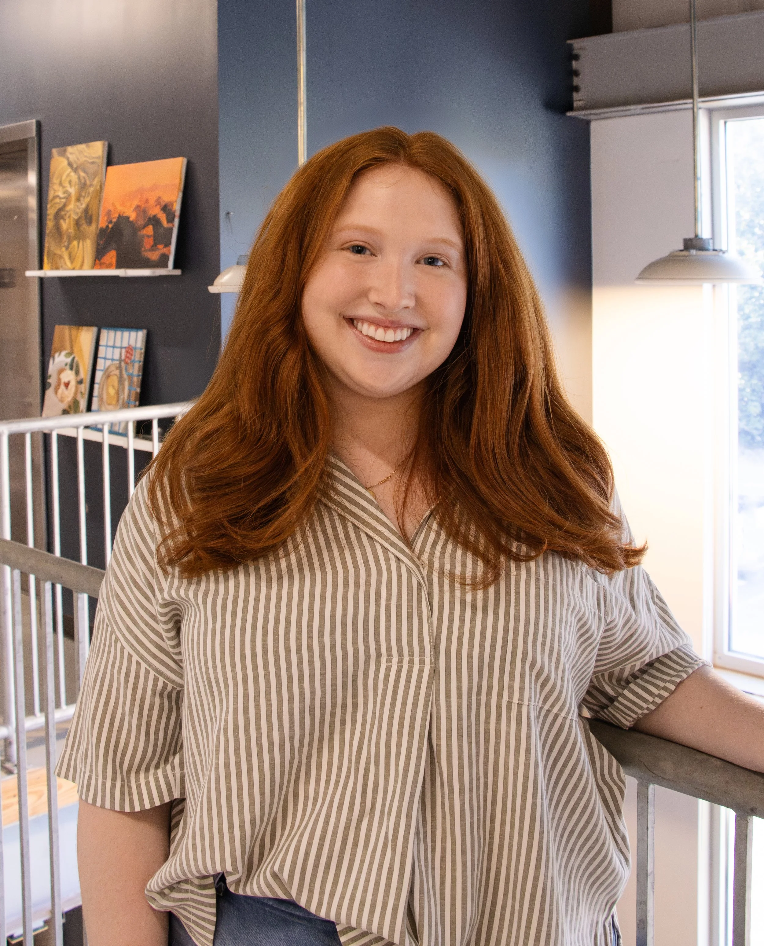 A young woman with long red hair, smiling, wearing a beige and white striped shirt, standing indoors near a railing, with paintings on a dark wall and a window in the background.