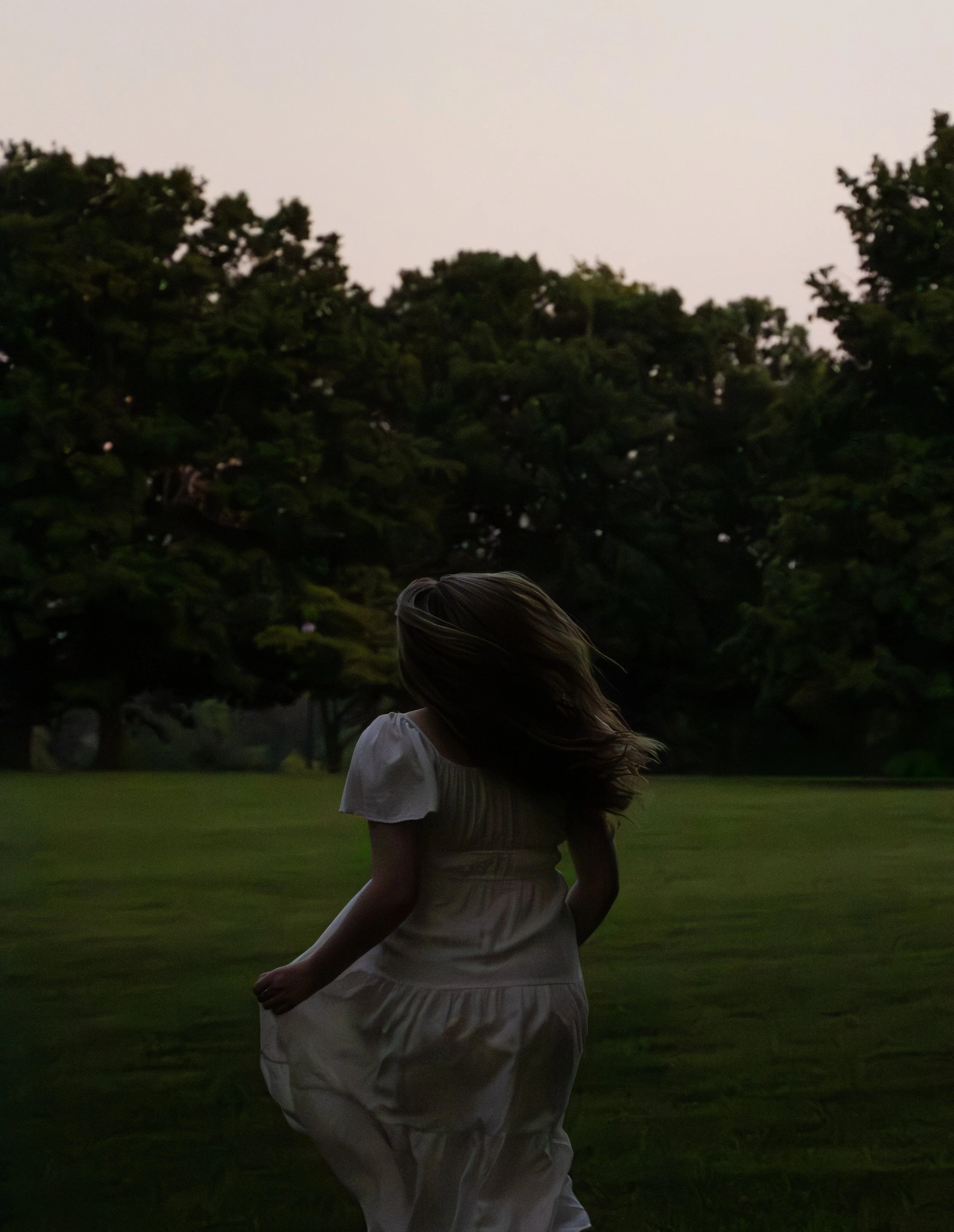 A girl in a white dress walking in a grassy field at dusk with trees in the background