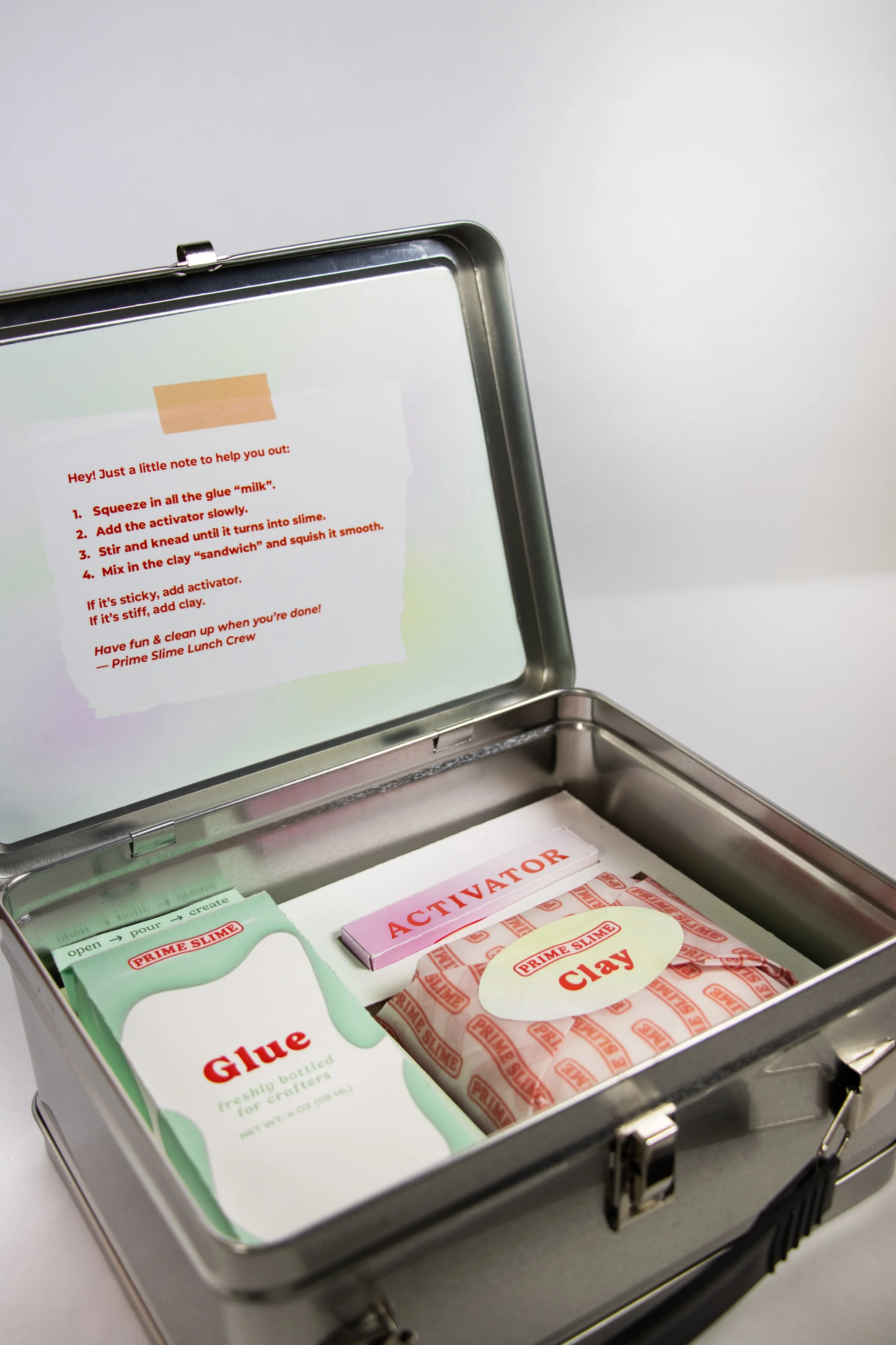 A metal box containing slime-making supplies, including glue, activator, and clay, with instructions displayed on the inside of the lid.