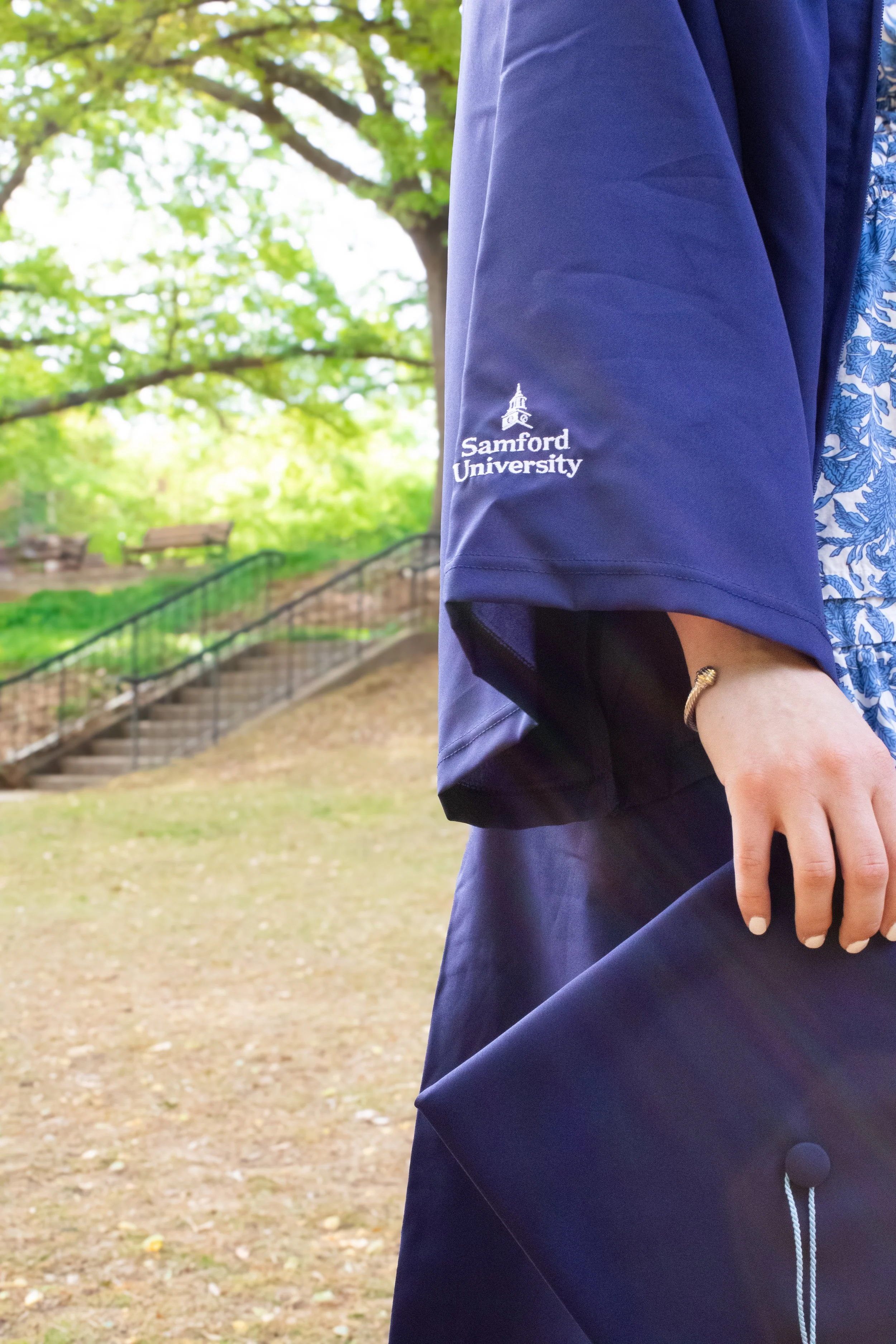 Close-up of a person wearing a blue Samford University graduation gown and holding a diploma, outdoors with trees and a staircase in the background.