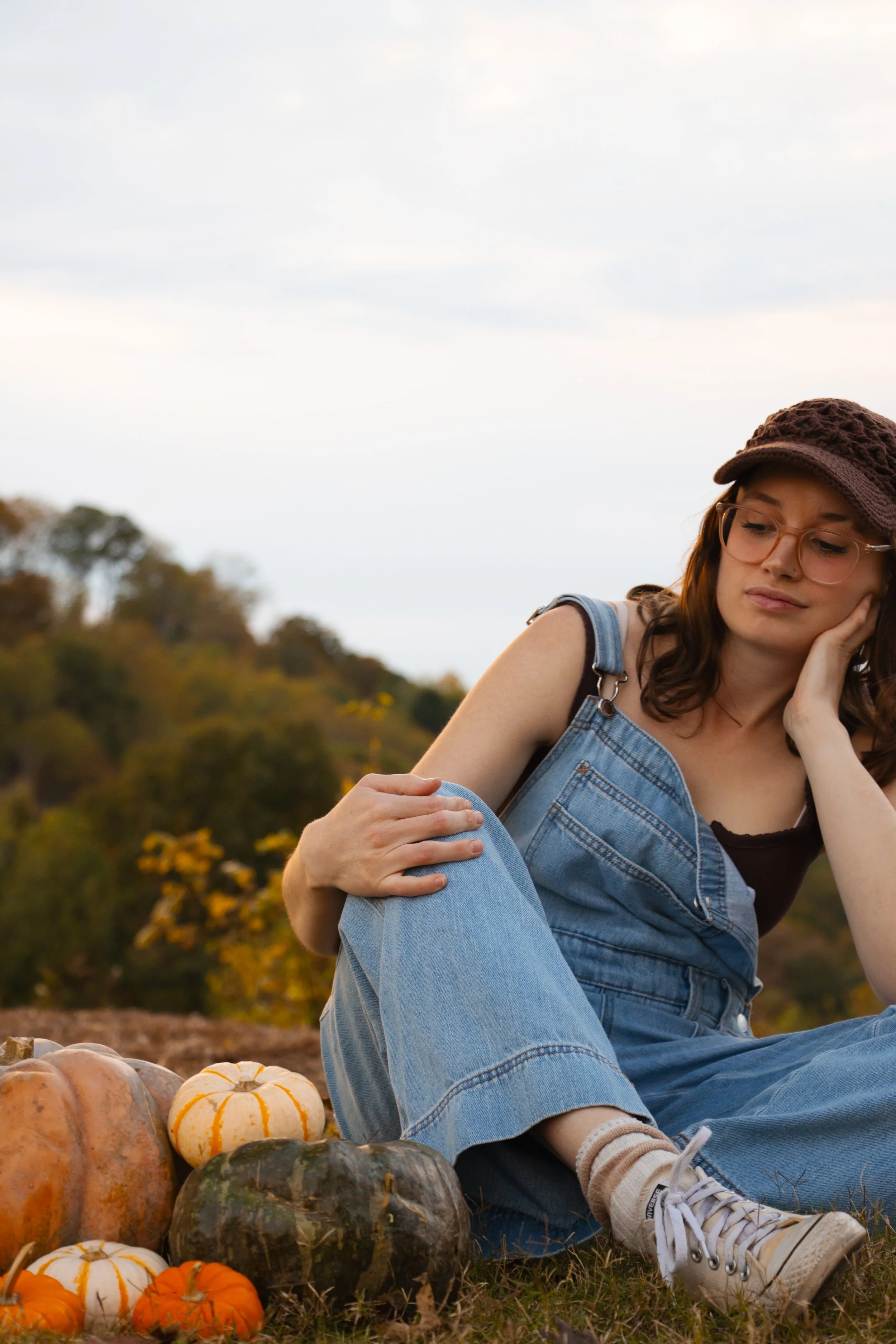 A young woman sitting outdoors among pumpkins, wearing denim overalls, a brown cap, glasses, and sneakers, resting her head on her hand with a pensive expression.