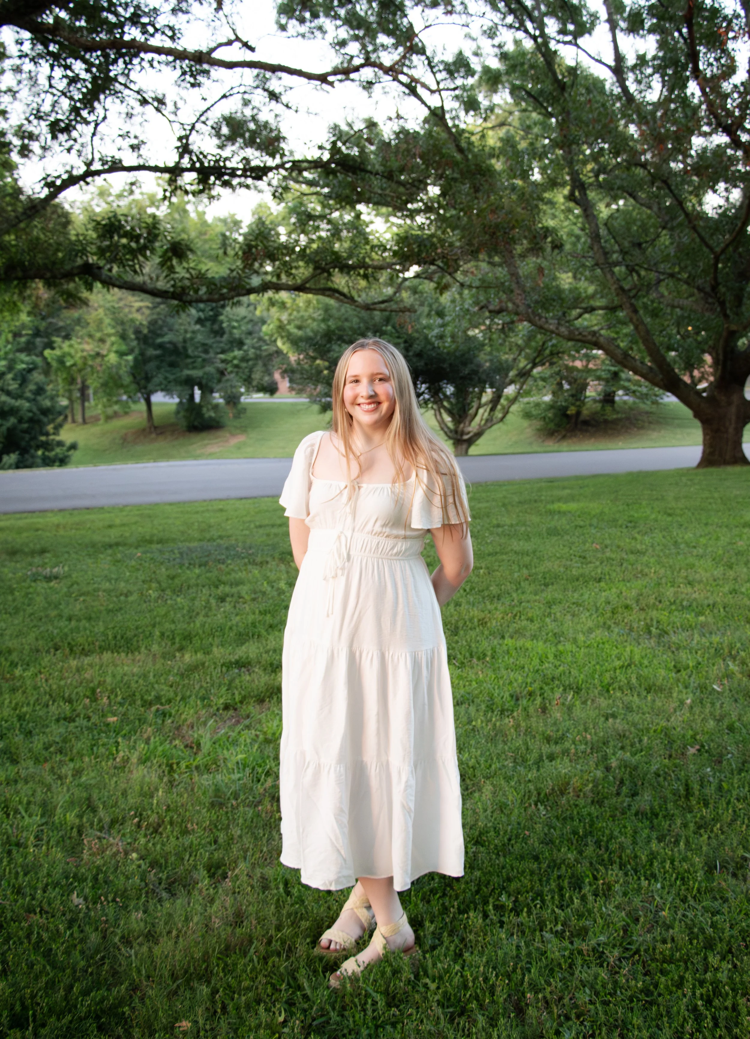 A young woman with long blonde hair, wearing a white dress, stands on a grassy area under trees, smiling at the camera.