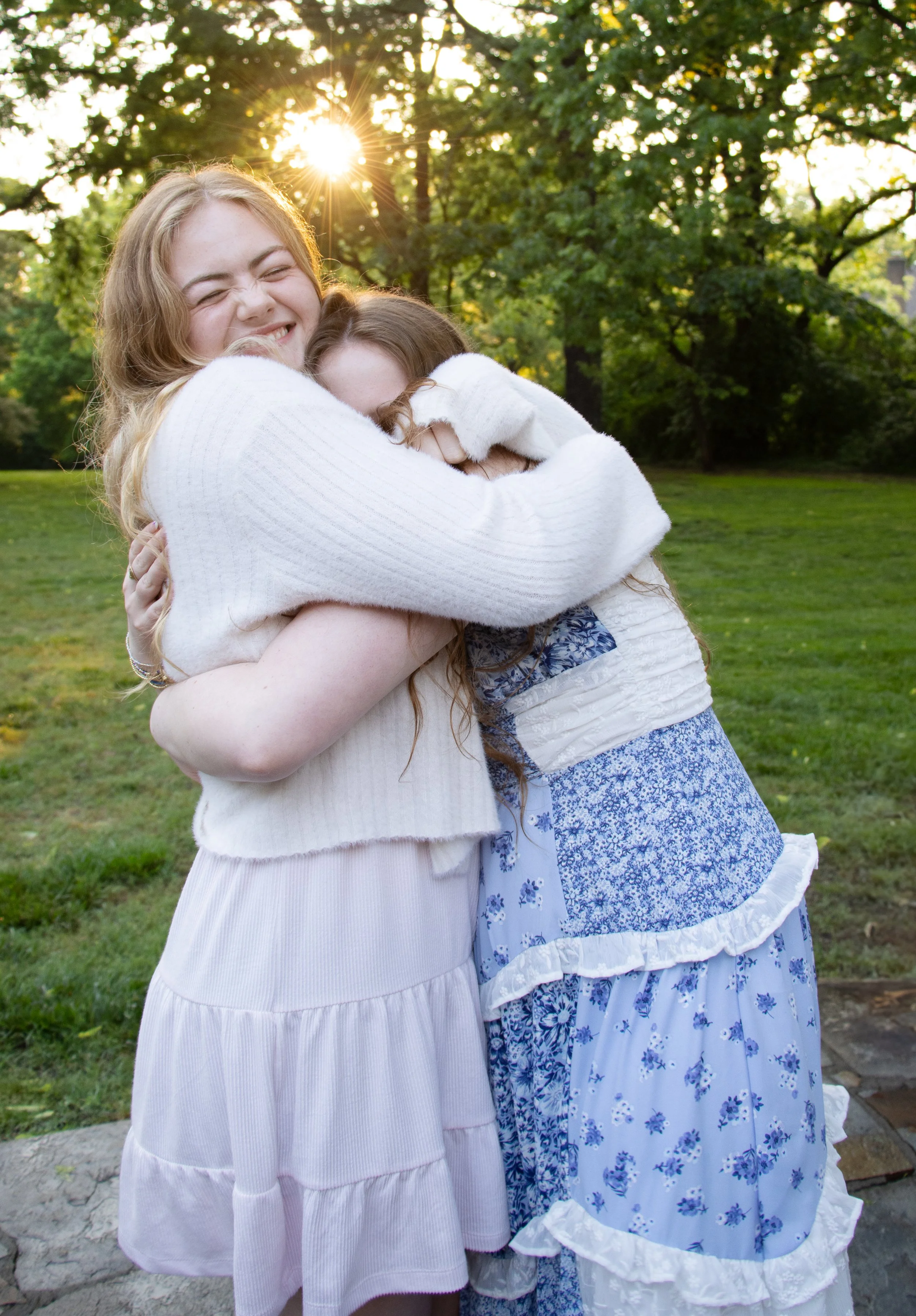 Two women hugging each other in a park, with sunlight filtering through trees in the background.