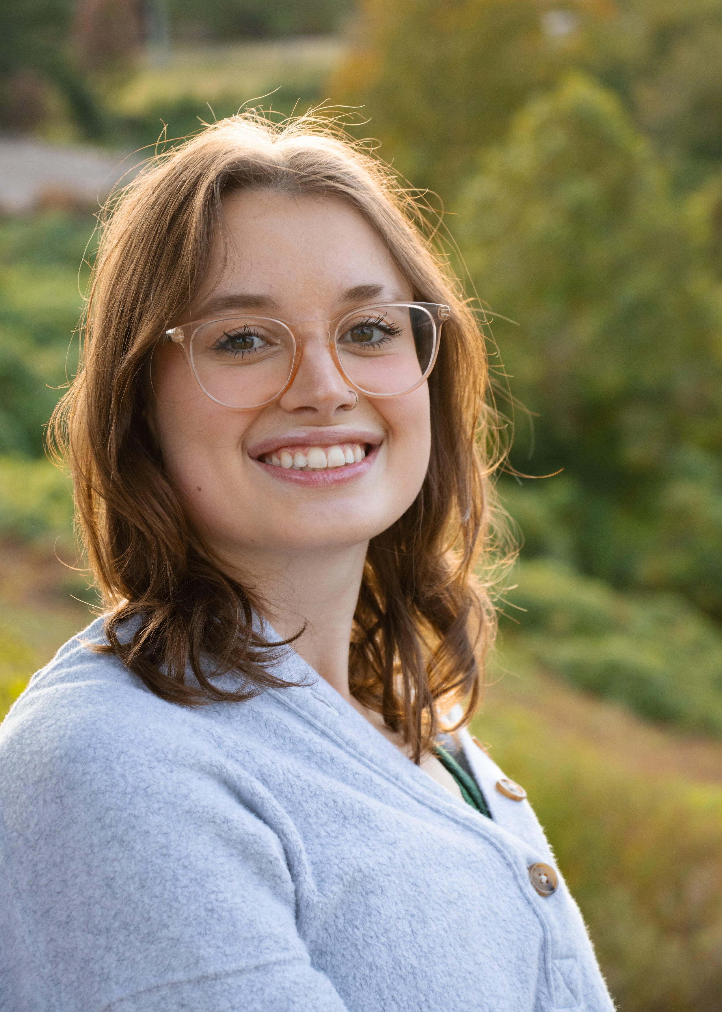 A young woman with glasses smiling outdoors in a park or garden with green trees and blurred background.