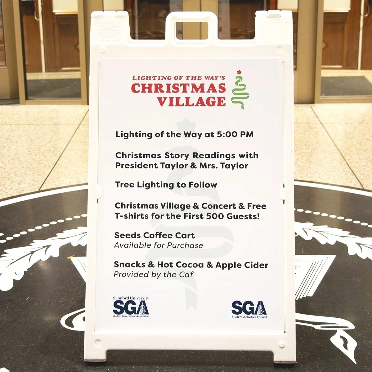Event signboard announcing Christmas village activities at Samarford University, including lighting of the way at 5:00 PM, story readings with the Taylors, tree lighting, free T-shirts for first 500 guests, a coffee cart, and snacks and hot cocoa.