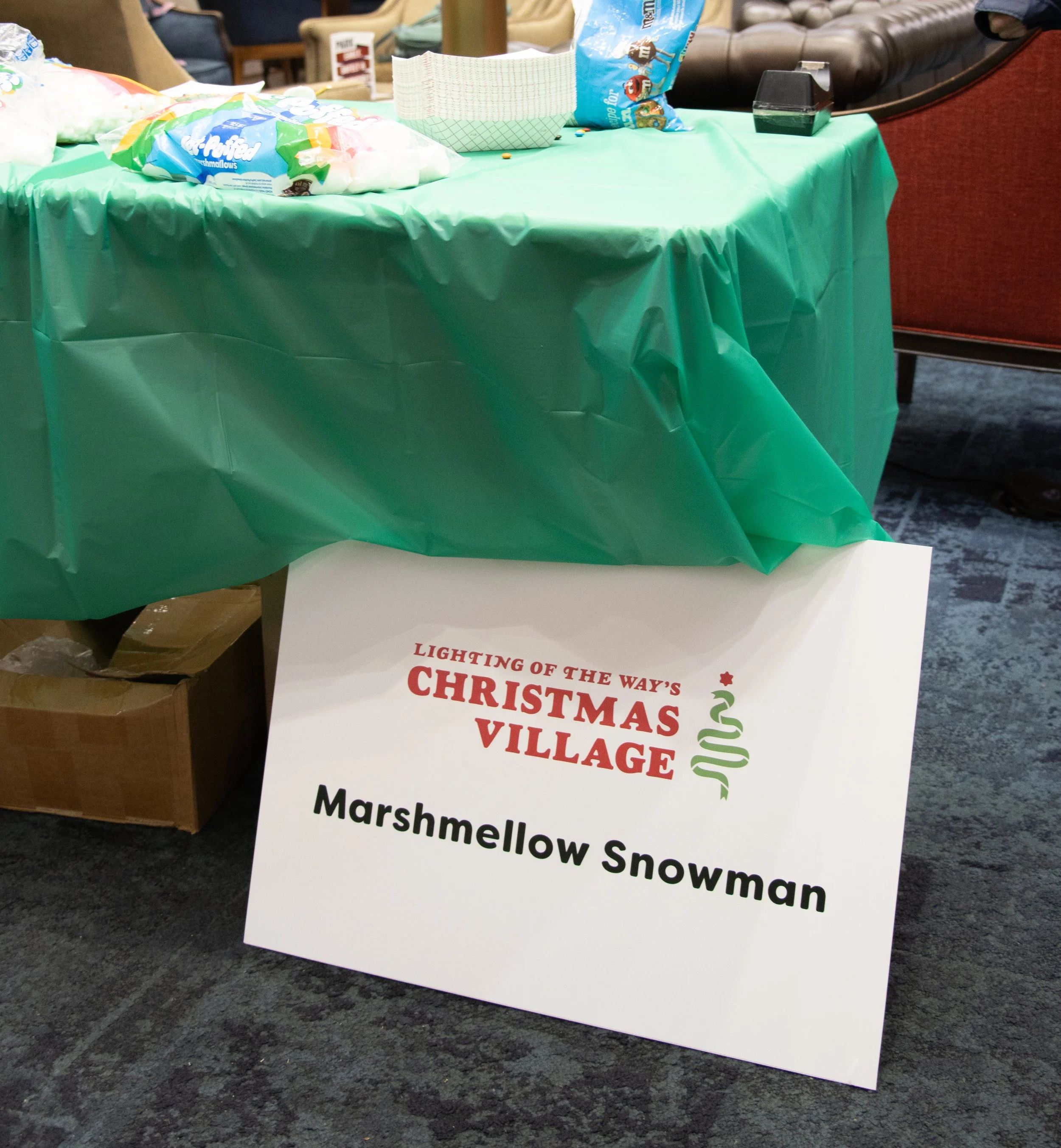 Sign reading "Lighting of the Way's Christmas Village, Marshmellow Snowman" placed on the floor in front of a table covered with a green tablecloth, with marshmallow treats and snack bags on top, in an indoor setting.