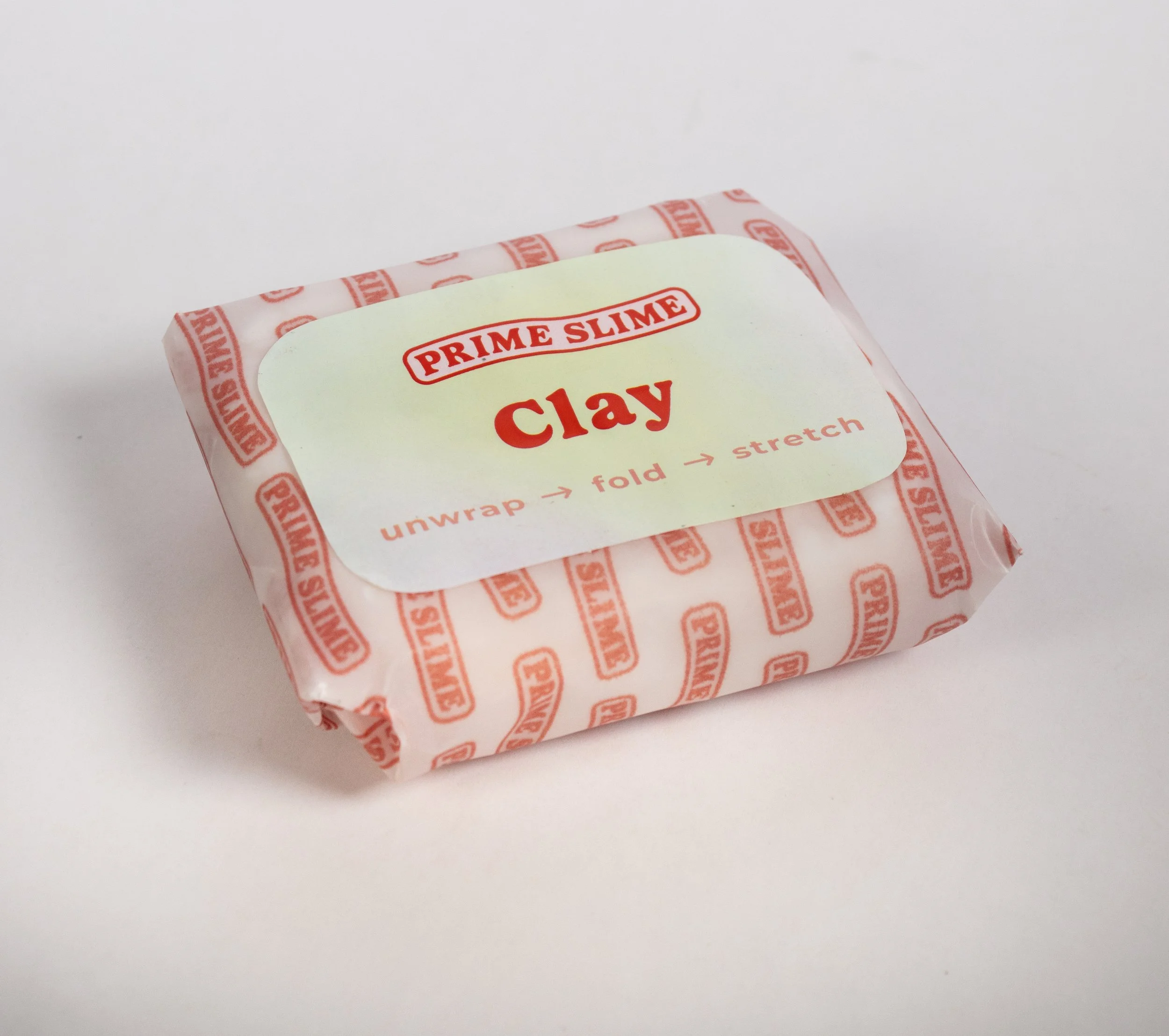 A small rectangular package of Prime Slime clay wrapped in white paper with red text and logo, resting on a plain white surface.