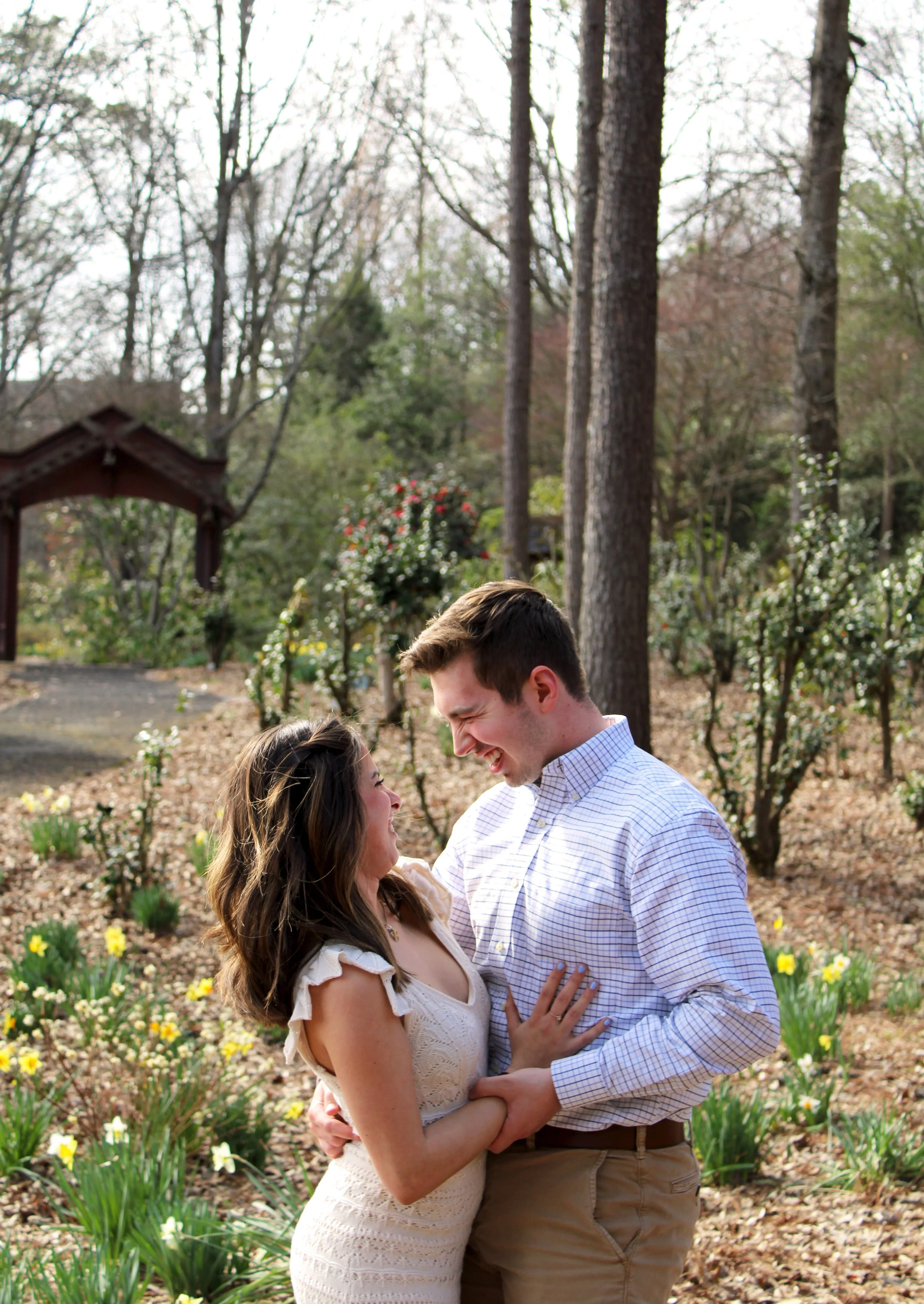 A young couple is embracing and laughing in a lush garden with blooming flowers and tall trees.
