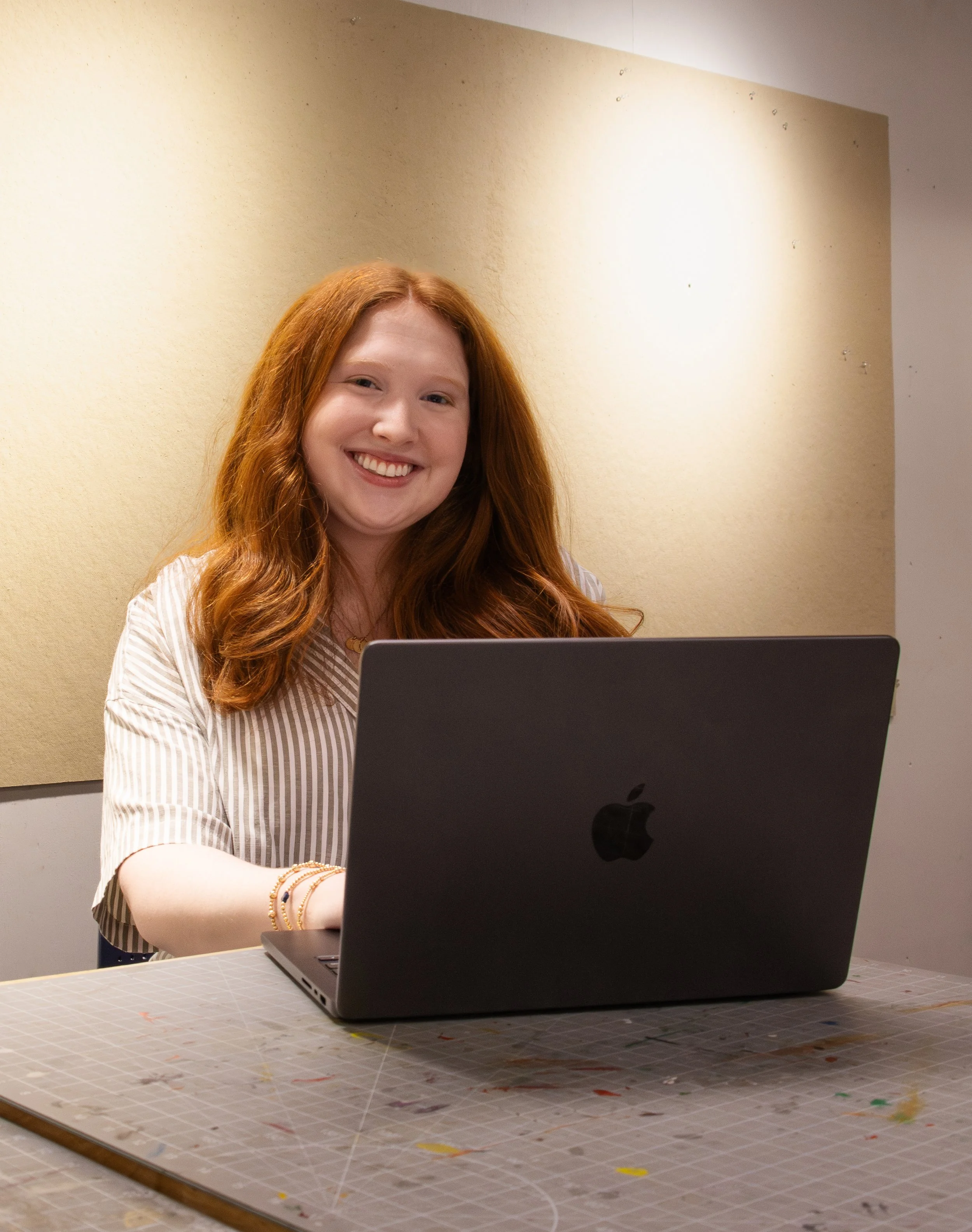 A young woman with red hair smiling while using a black MacBook laptop, sitting at a table with a gray surface, in an indoor setting with a beige wall in the background.