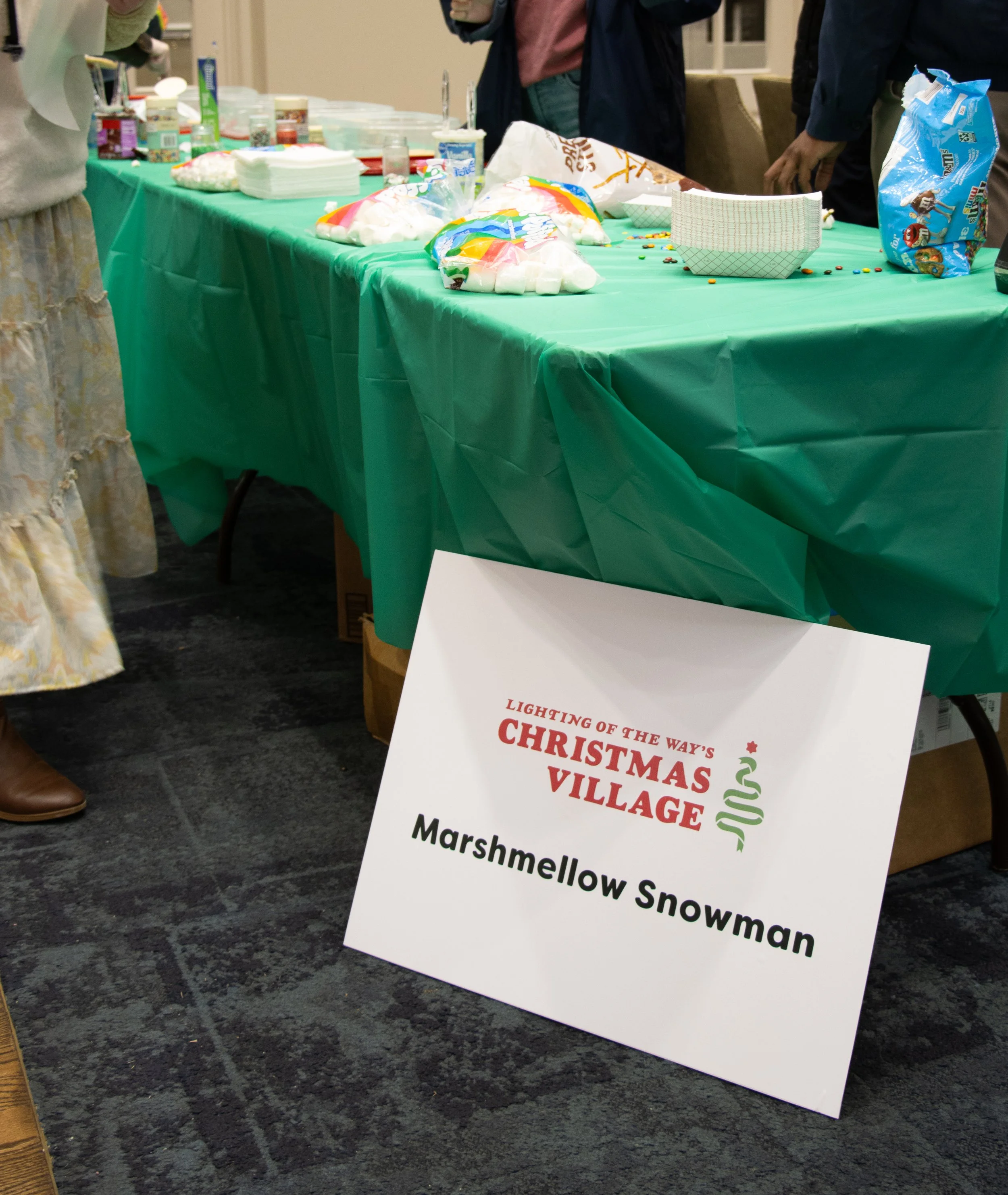 Table with green tablecloth holding marshmallow snowman supplies, marshmallows, and sweets, with people standing nearby, and a sign in front reading "Lighting of the Way's Christmas Village, Marshmallow Snowman."