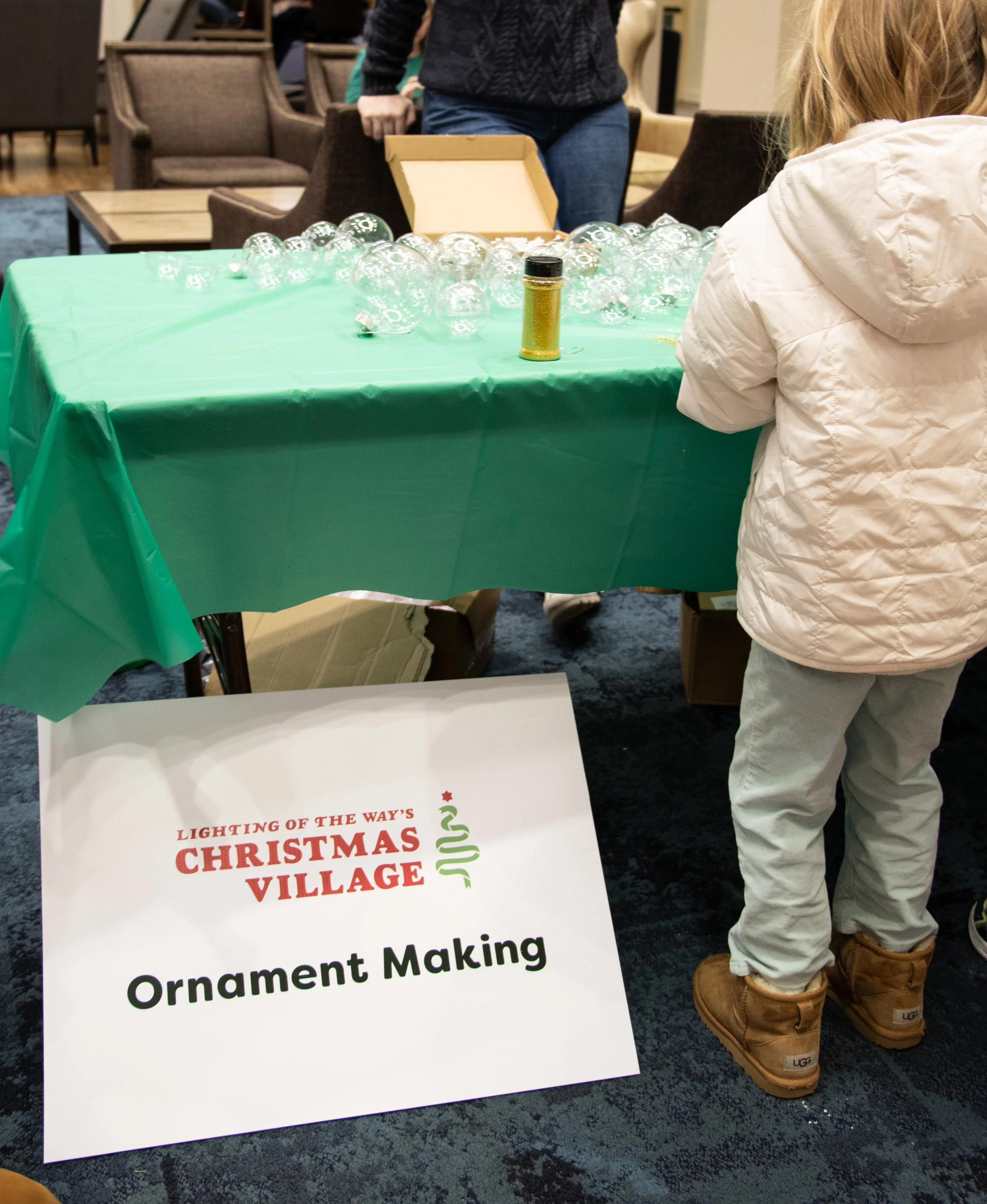 A table with a green tablecloth displaying clear Christmas ornaments and a gold glitter bottle at a Christmas village ornament making event, with a child in white coat and brown boots in foreground and people in background.