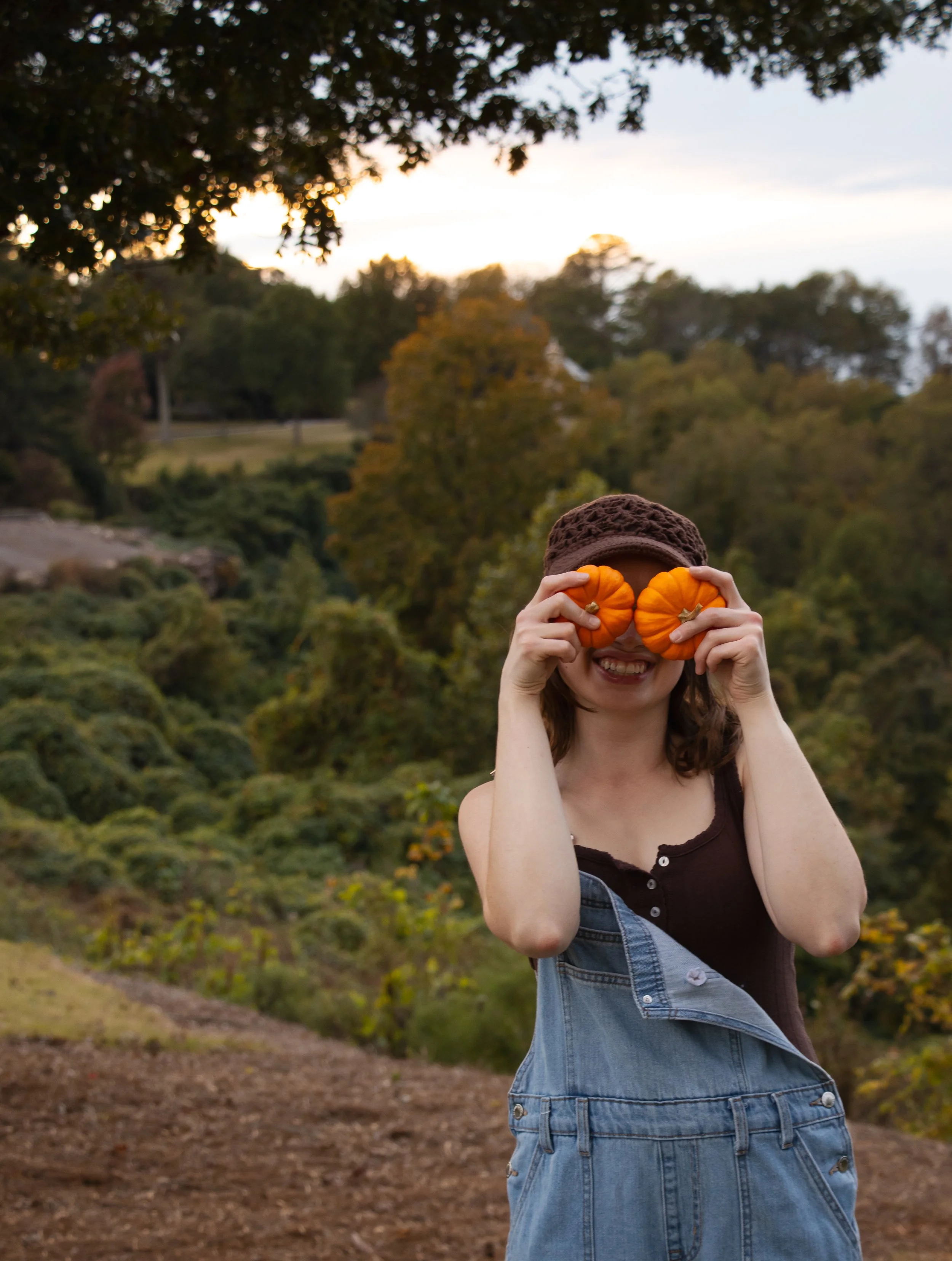 A woman standing outdoors in a park, holding small pumpkins over her eyes, smiling, with trees and a cloudy sky in the background.