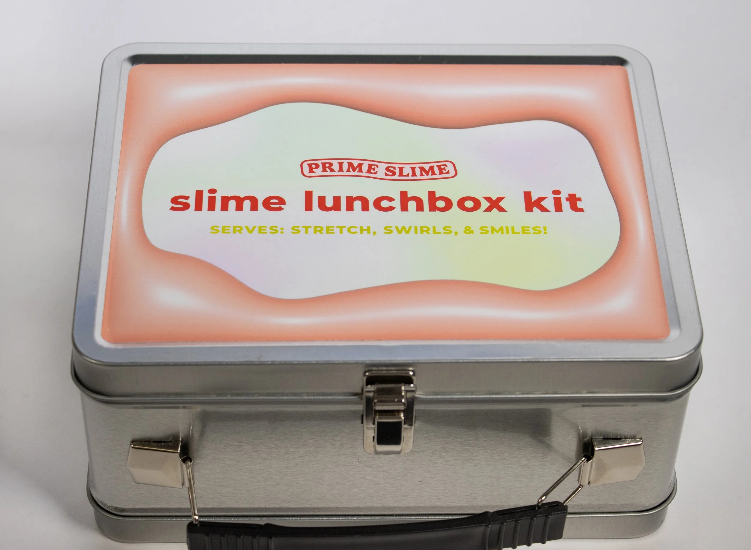 A metal lunchbox with a colorful label on the lid that reads "Prime Slime slime lunchbox kit, serves: stretch, swirls, & smiles!"
