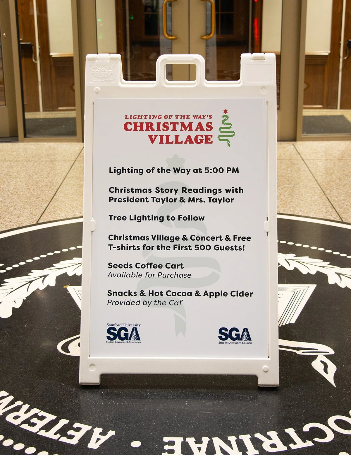 A white A-frame sign in front of a building entrance advertising the Christmas Village event, featuring Christmas story readings, tree lighting, free T-shirts, coffee cart, snacks, and apple cider, with Samford University Student Government Associati