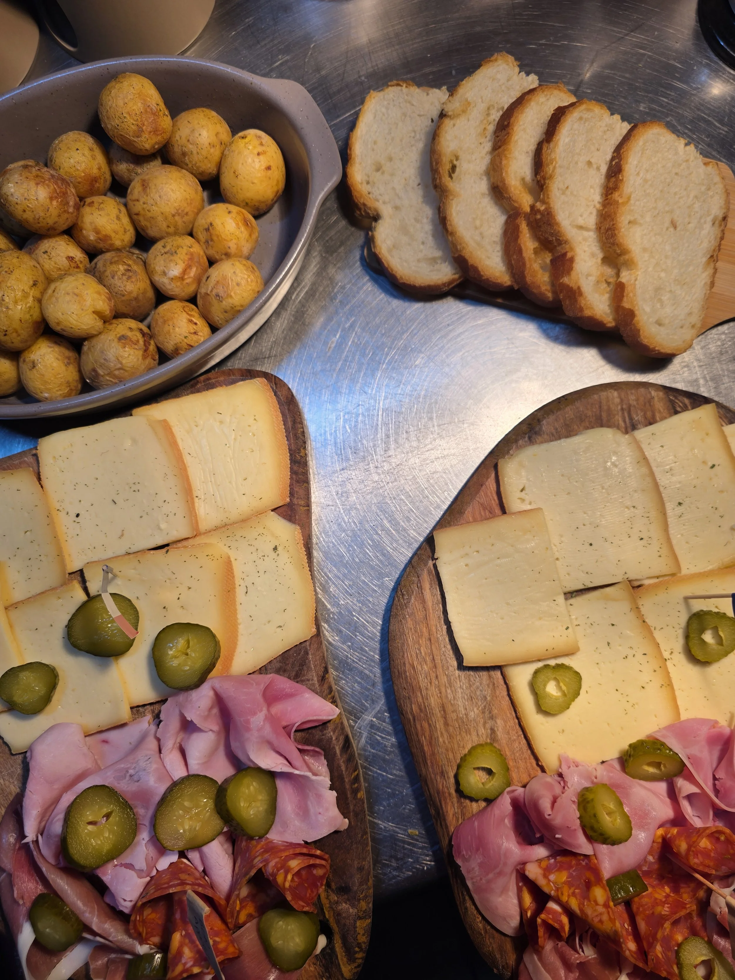 Assorted cheeses, sliced bread, small potatoes, and an assortment of meats with pickles on wooden and metal surfaces
