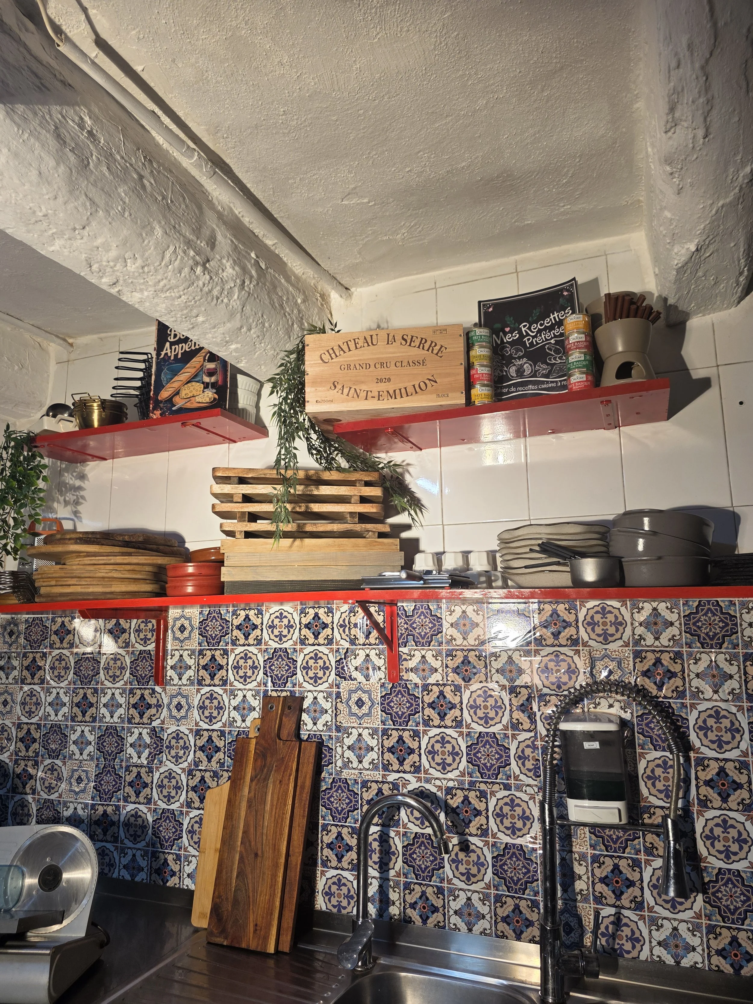 Kitchen with patterned tile backsplash, wooden cutting boards, stacked plates, and shelves with pots, cans, and decorative items.