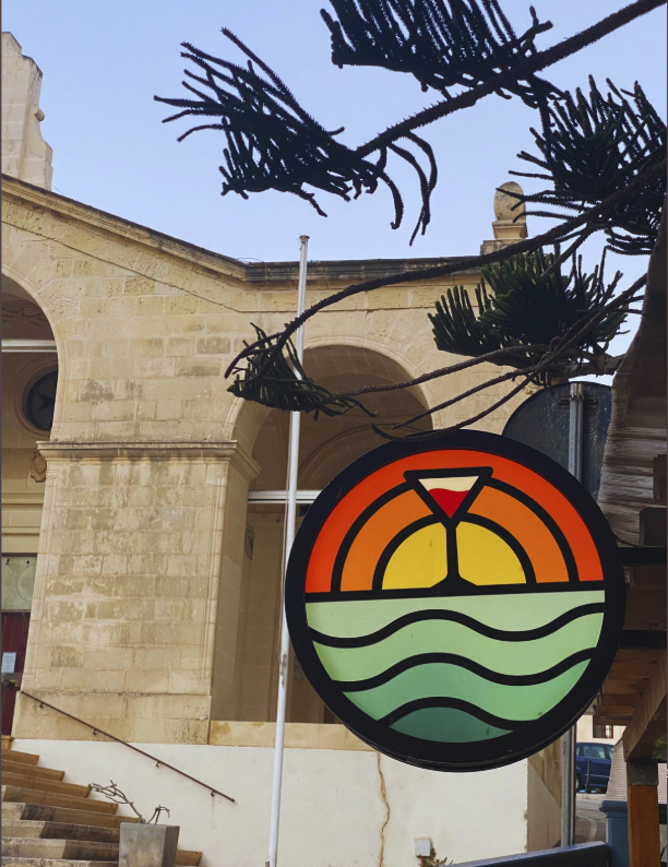 A circular sign with a sunset, water, and a cocktail glass, hanging outside a building with stone architecture and some tree branches overhead.
