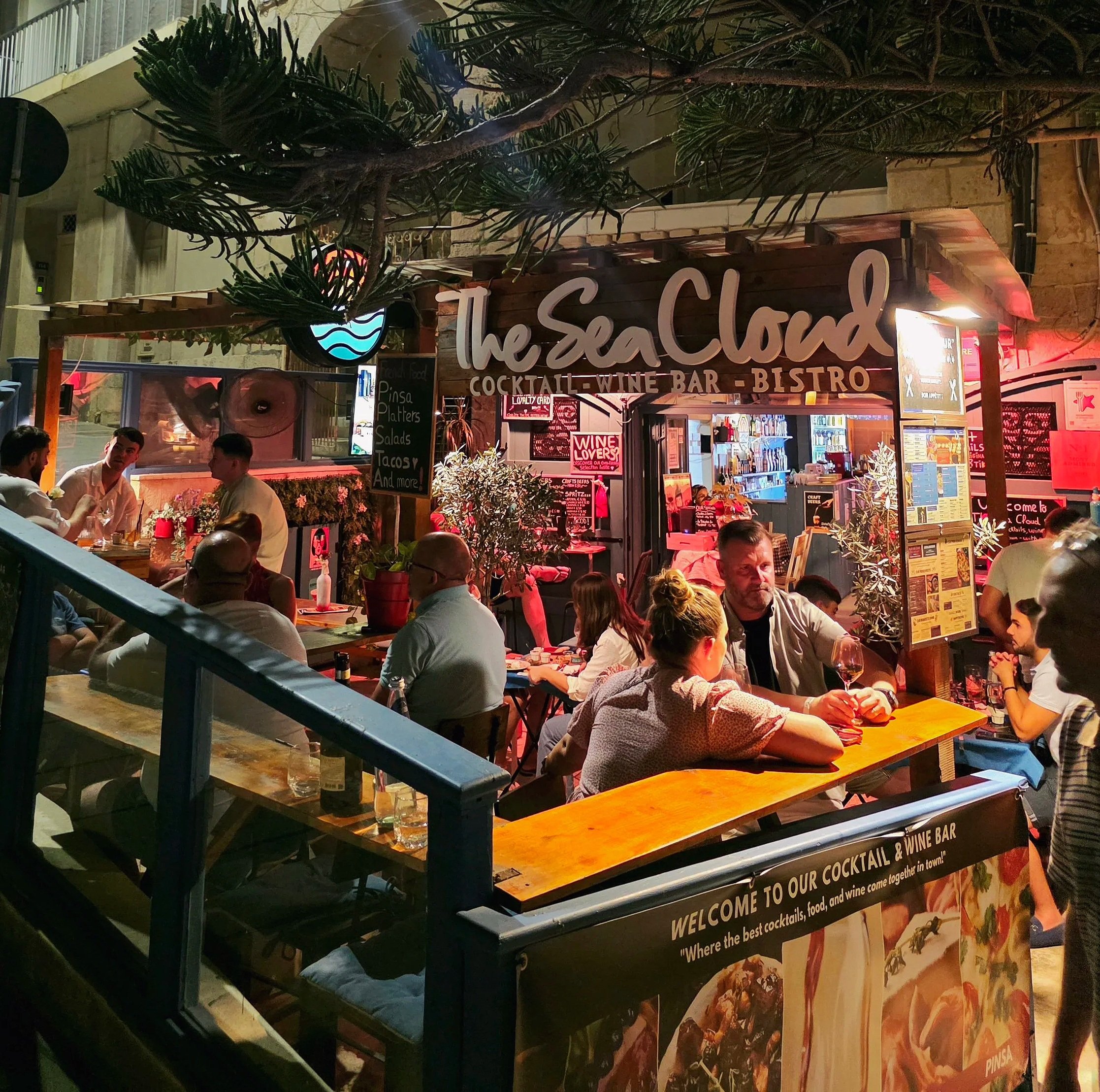 People dining outside at The Sea Cloud cocktail and wine bar bistro at night, with neon and warm lighting, surrounded by potted plants and a wooden signboard.