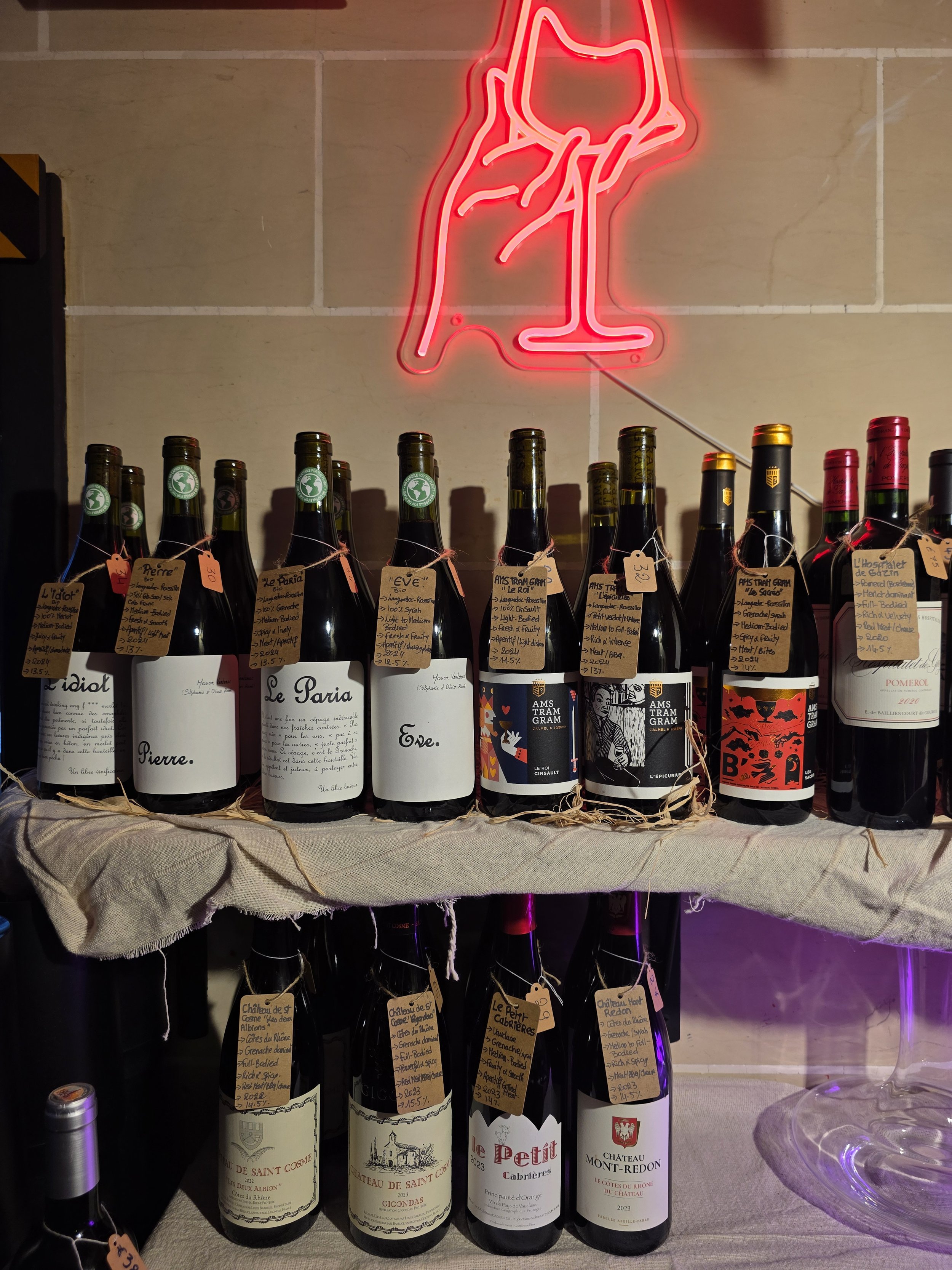 Display of wine bottles with handwritten tags, illuminated by a red neon sign of a hand holding an drink, against a beige tiled wall.