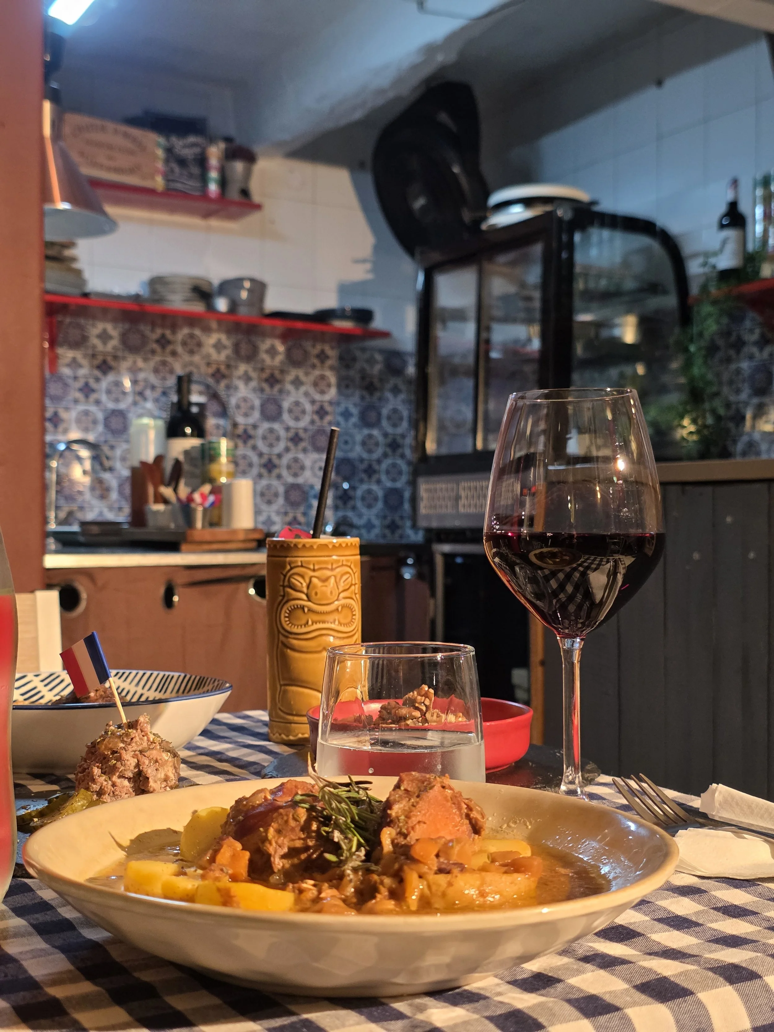 A bowl of pasta with sauce, a glass of red wine, and a glass of water on a checkered tablecloth in a cozy restaurant kitchen setting.