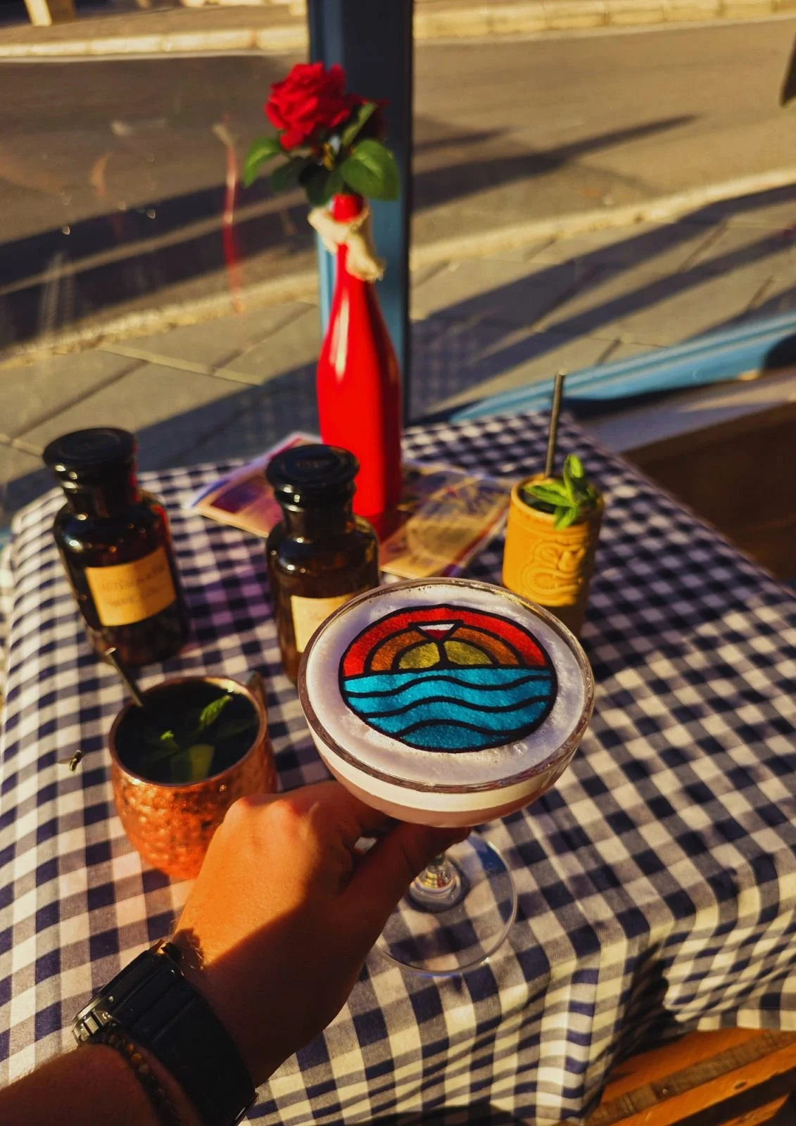 A hand holding a cocktail with a colorful sunset and ocean design on top, in front of a table with bottles, a copper mug with mint, and a red flower in a red vase.