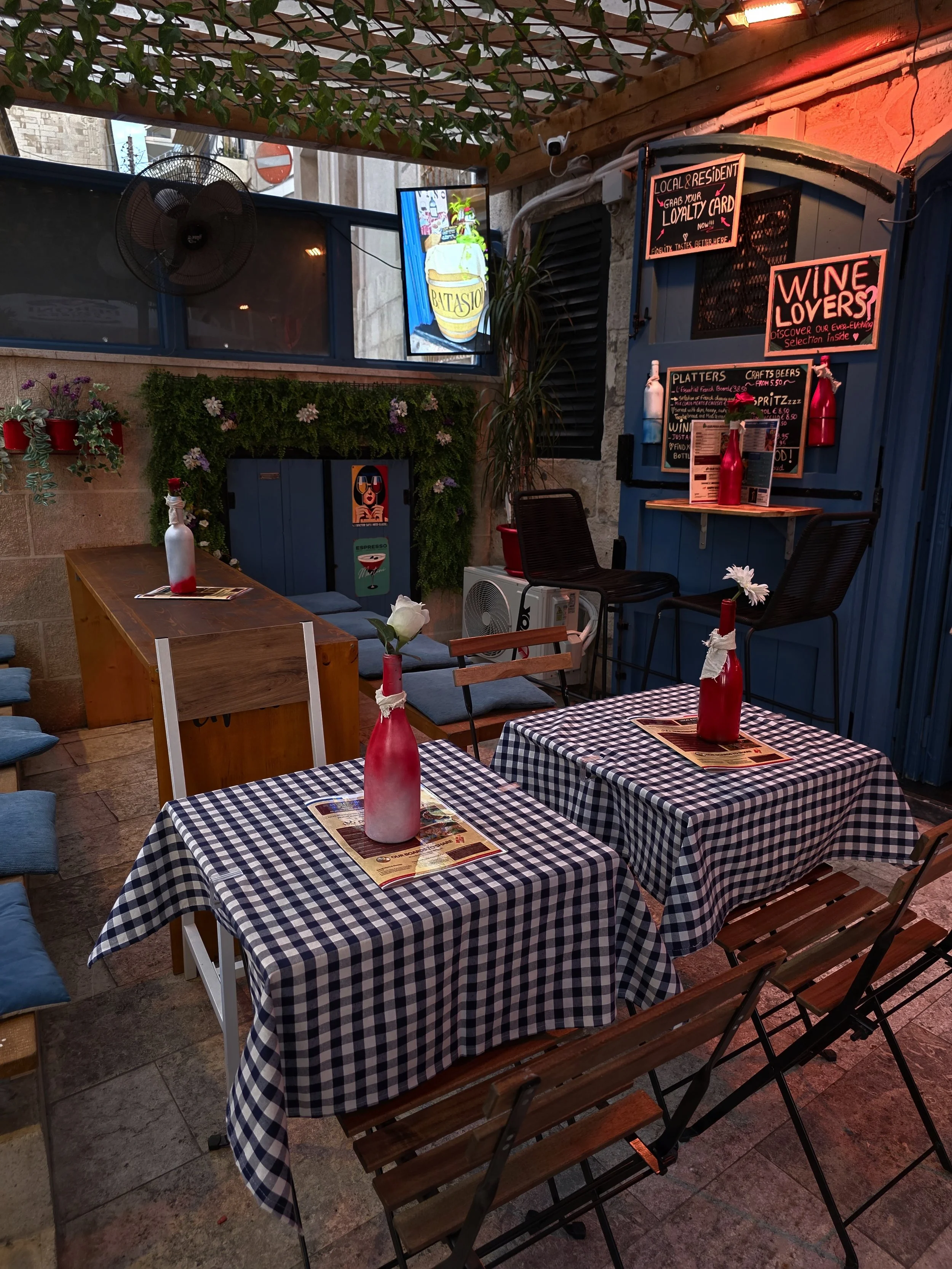 An outdoor cafe seating area with two tables covered in blue and white checkered tablecloths, each with a red bottle holding a white flower. There are wooden and metal chairs, some with blue cushions. The area is decorated with potted plants, pink fl