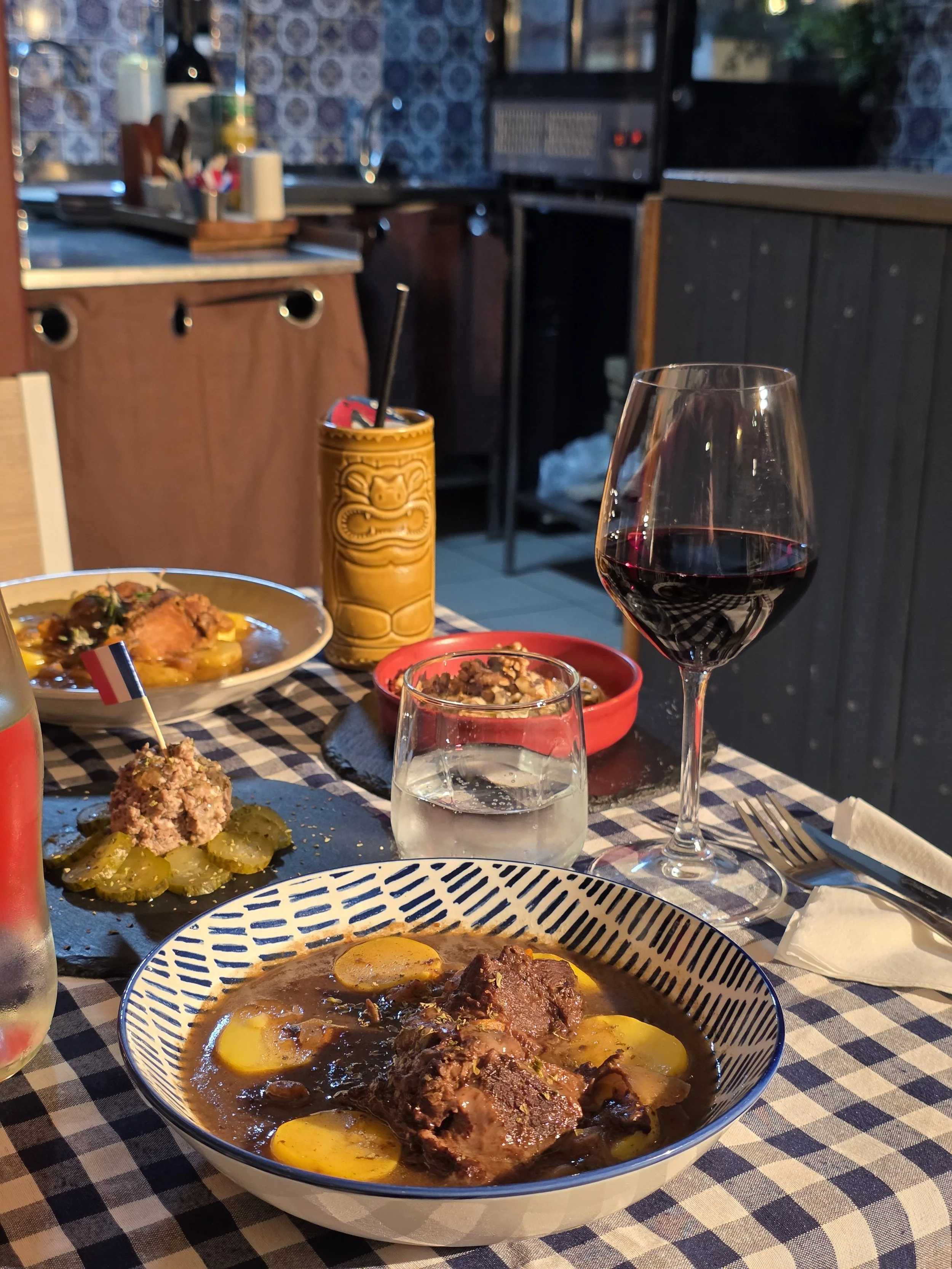 A dining table with various dishes, including a stew with meat and eggs in a patterned bowl, a plate of pickles with a meatball and flag toothpick, a glass of red wine, a glass of water, a yellow tiki mug, and additional plates and utensils, with a c