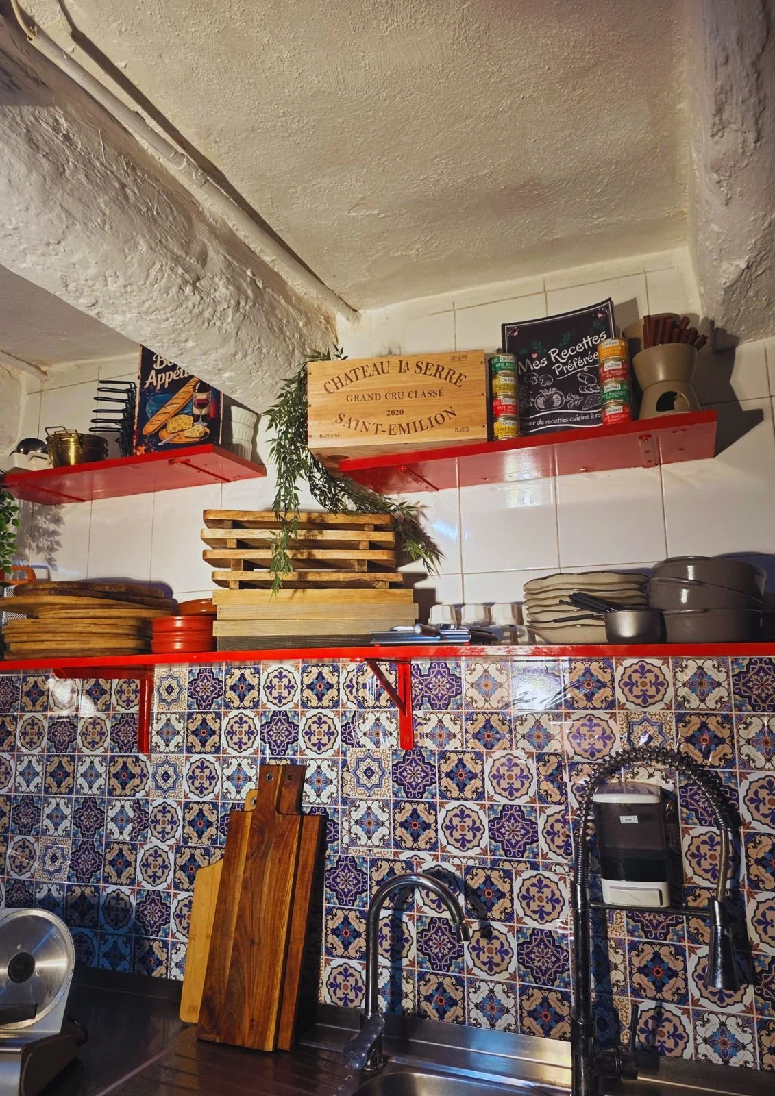 A kitchen shelf with wooden cutting boards, stacked plates, and spice jars. The wall behind has patterned tiles, and below is a stainless steel sink with a faucet.