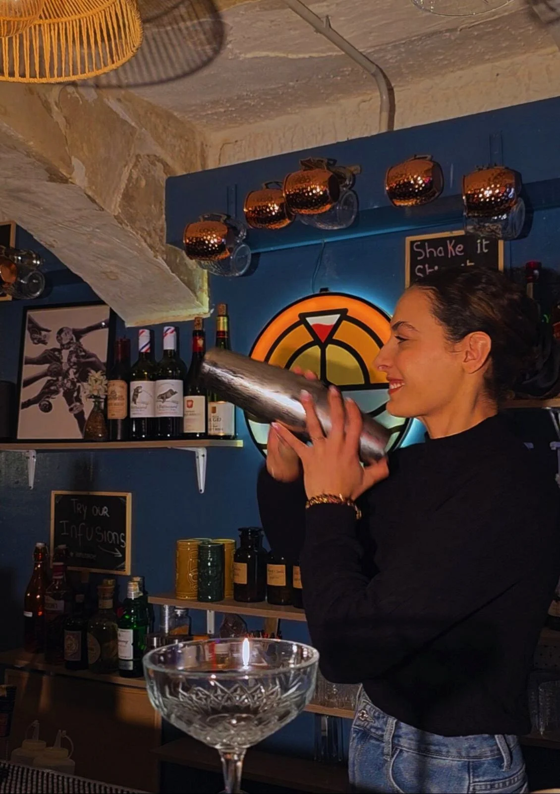 A woman is holding and preparing to drink from a cocktail shaker behind a bar with bottles of alcohol, barware, and decorative items, inside a dimly lit bar or restaurant.
