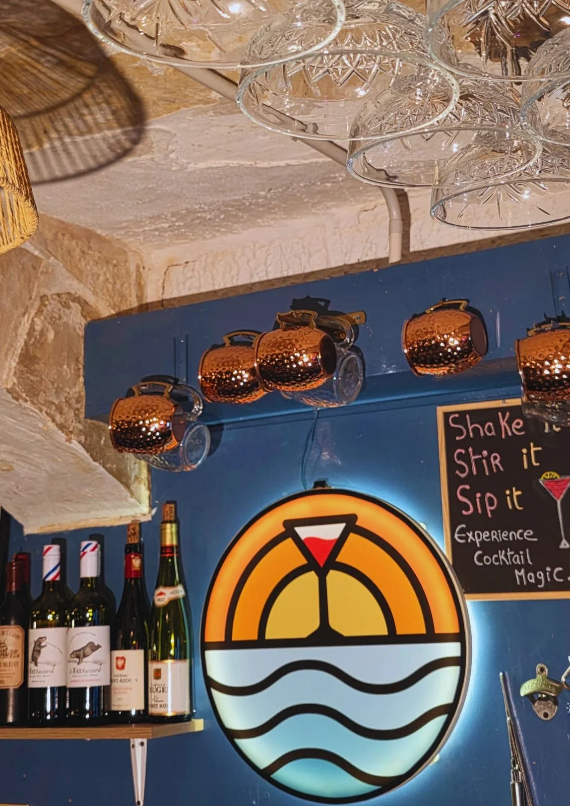 Decorative bar area with hanging glassware, copper mugs, liquor bottles, a sign with a drink recipe, and a circular illuminated artwork of a cocktail glass with waves and a red umbrella.
